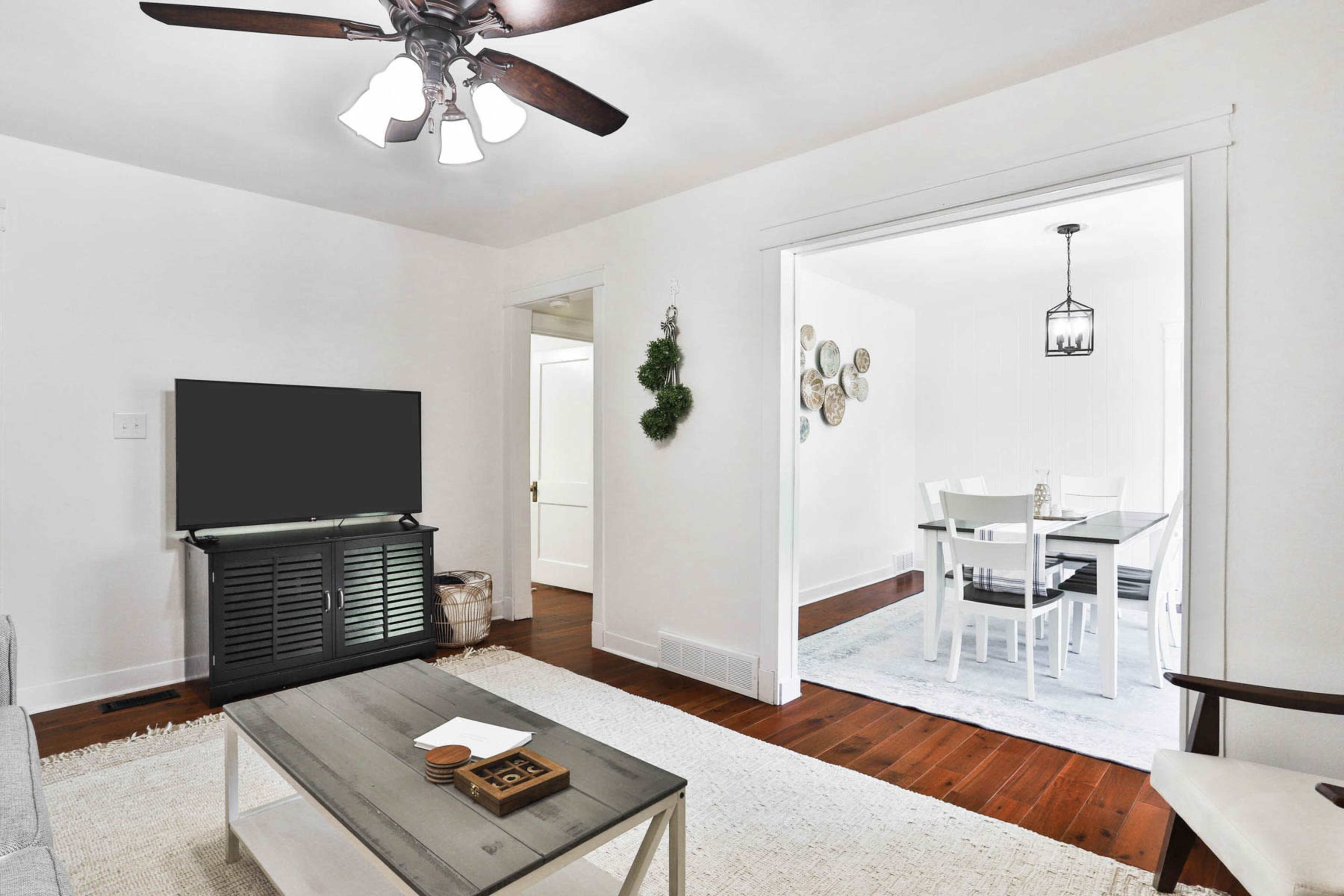 A living room features a ceiling fan, a television on a stand, and a table, with a doorway leading to a light-filled dining area.