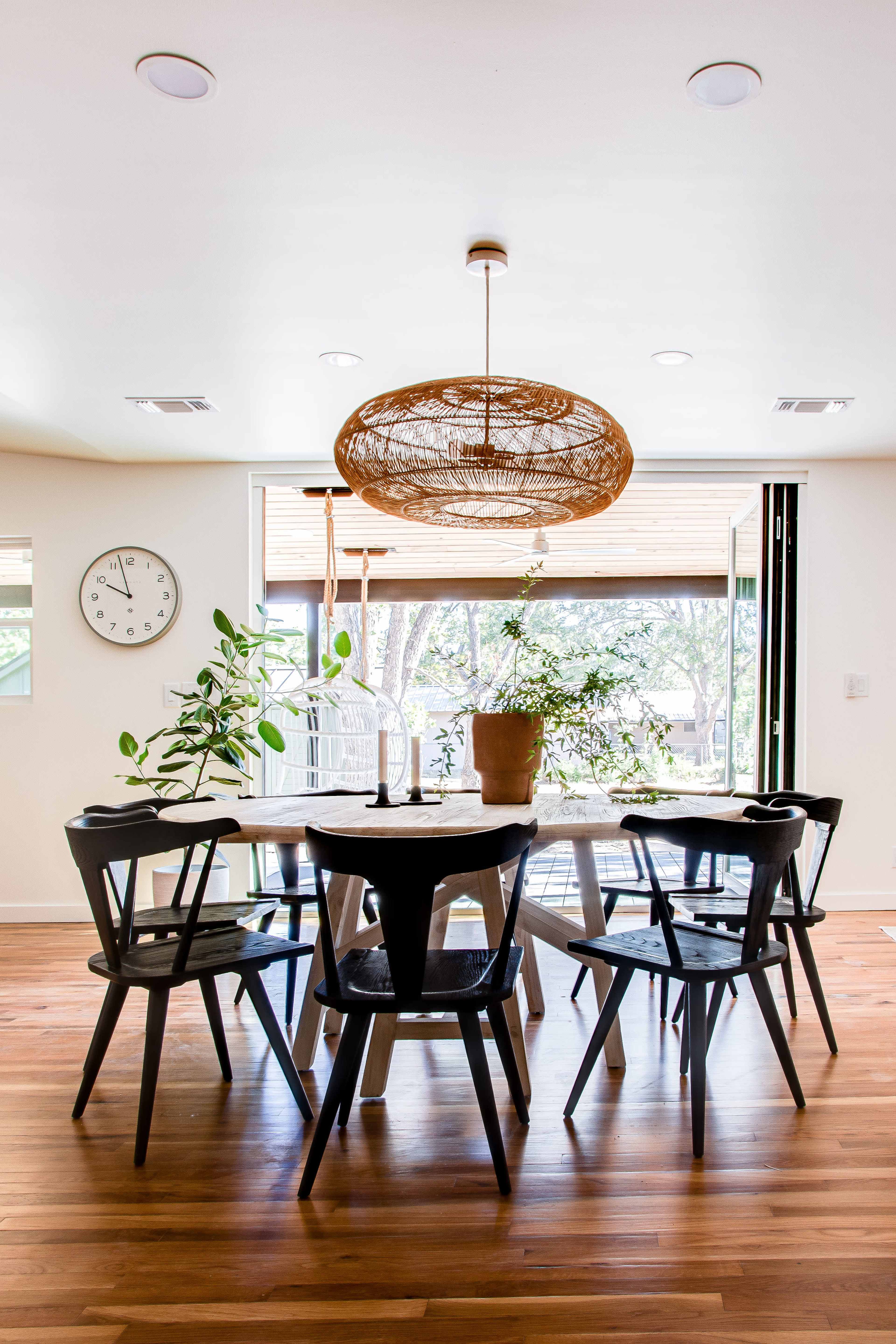A dining area features a round table surrounded by black chairs, with a large woven pendant light and potted plants nearby.