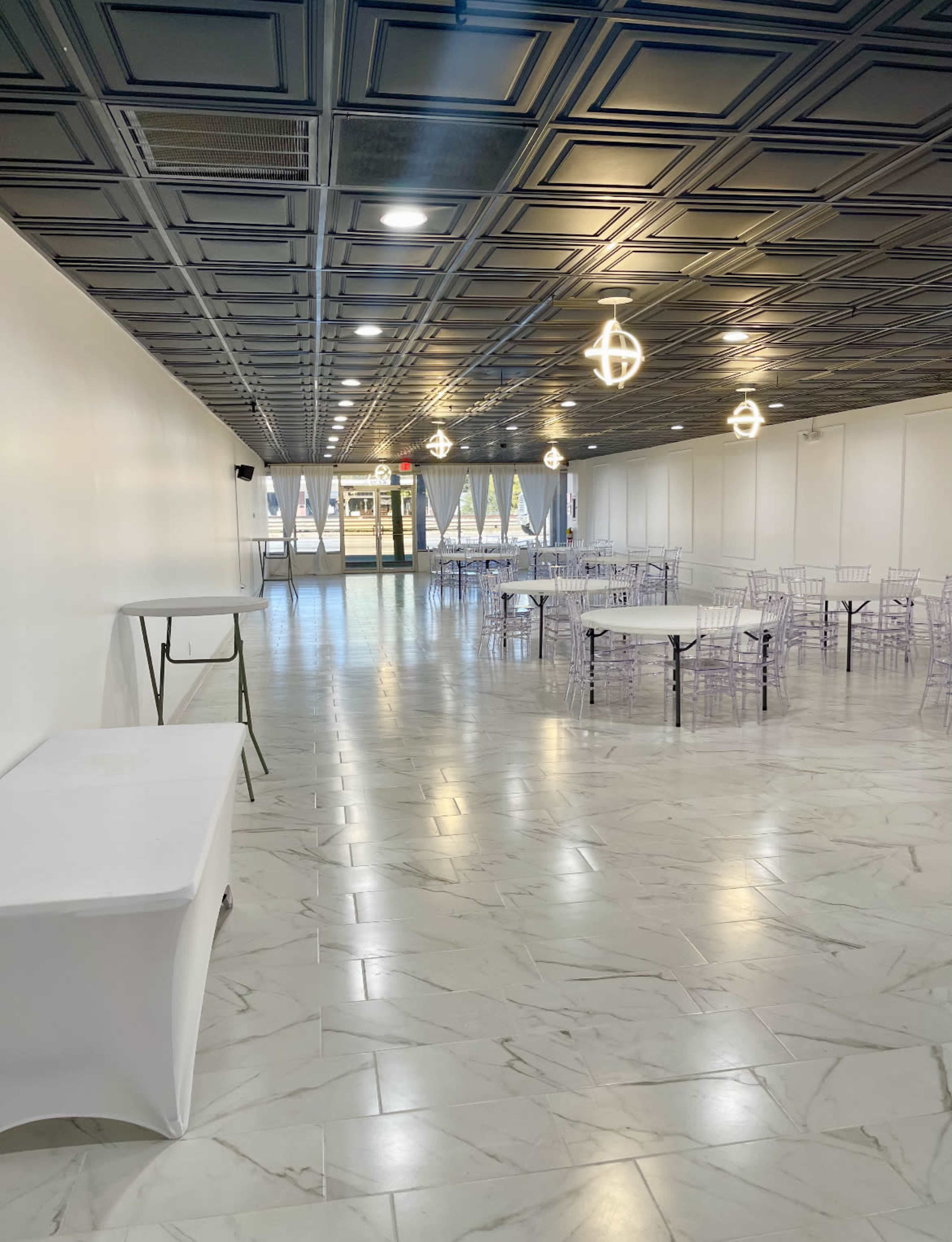 A spacious, well-lit event hall with multiple rectangular tables and transparent chairs arranged for a gathering.