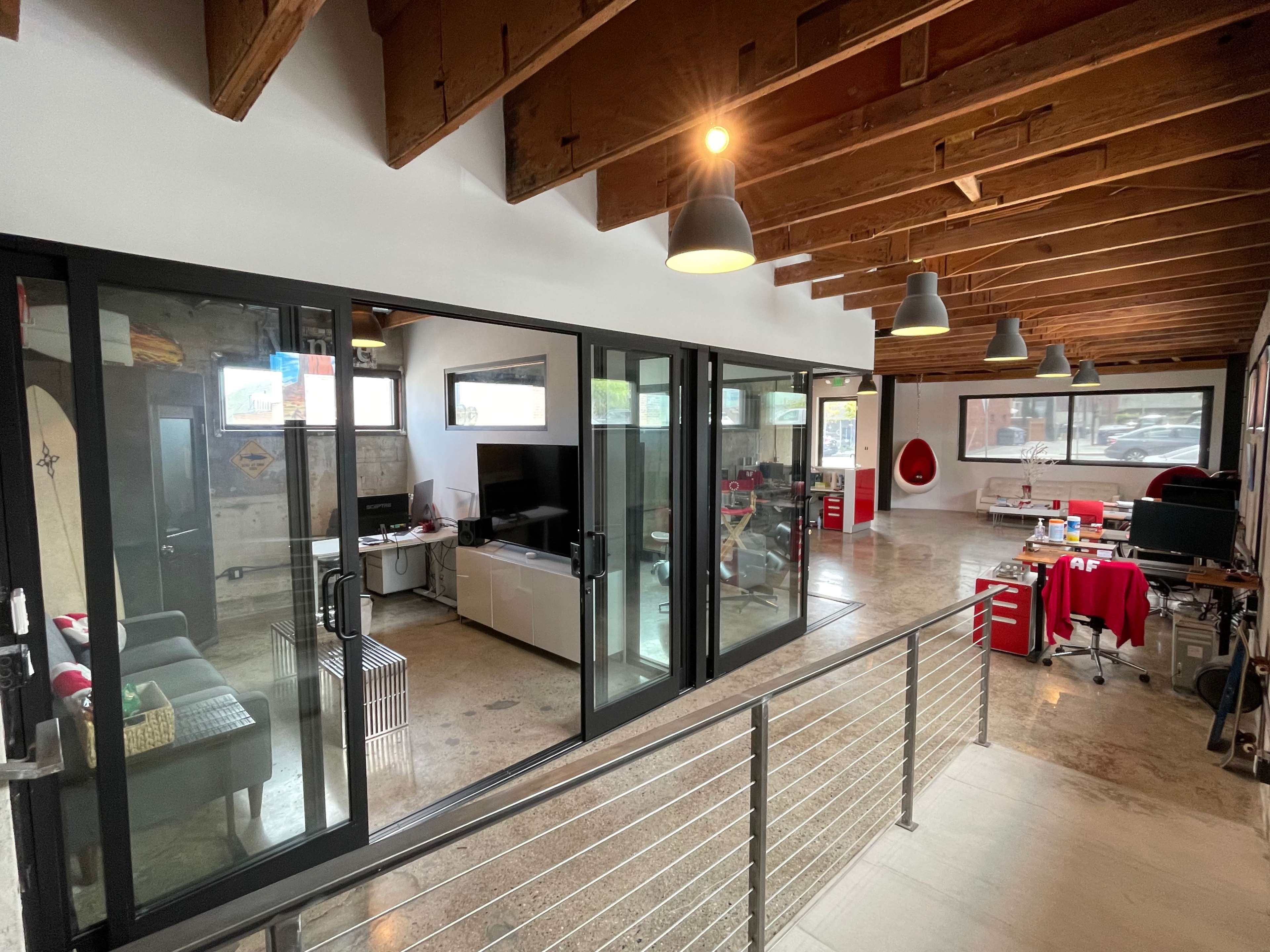 A modern office space with large glass walls, wooden beams above, and a mix of furniture including a couch, desks, and office equipment.