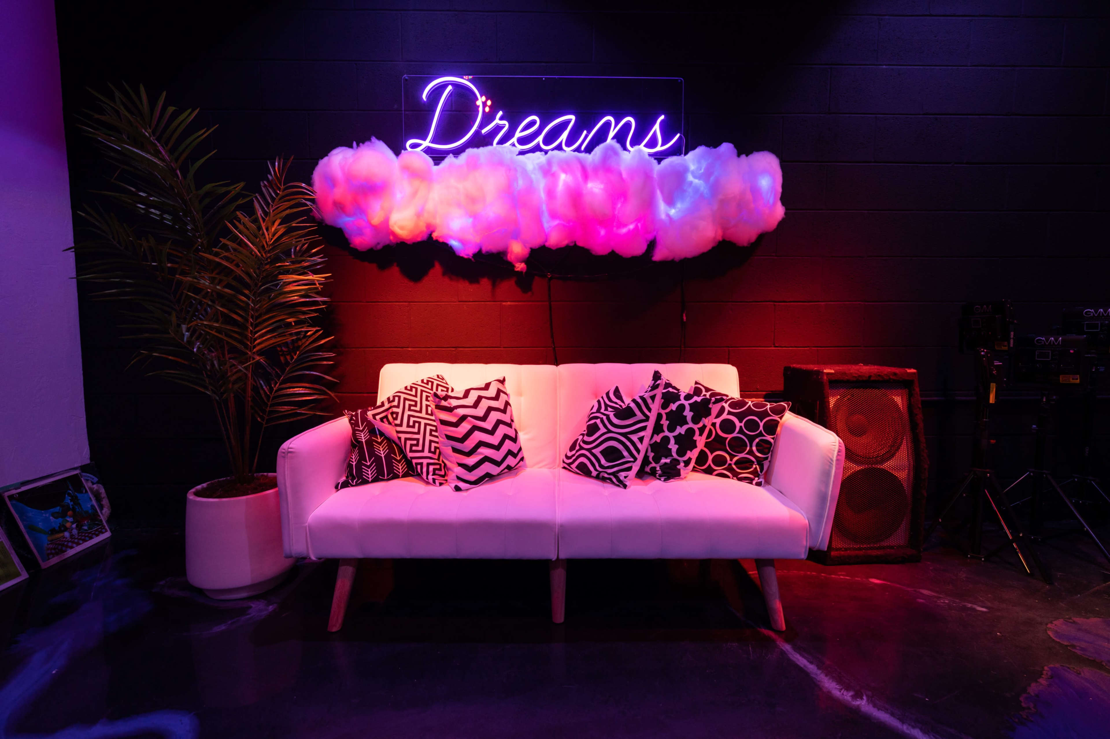 A white sofa with patterned pillows is positioned beneath a neon sign that says "Dreams," against a black wall adorned with decorative clouds and a plant.