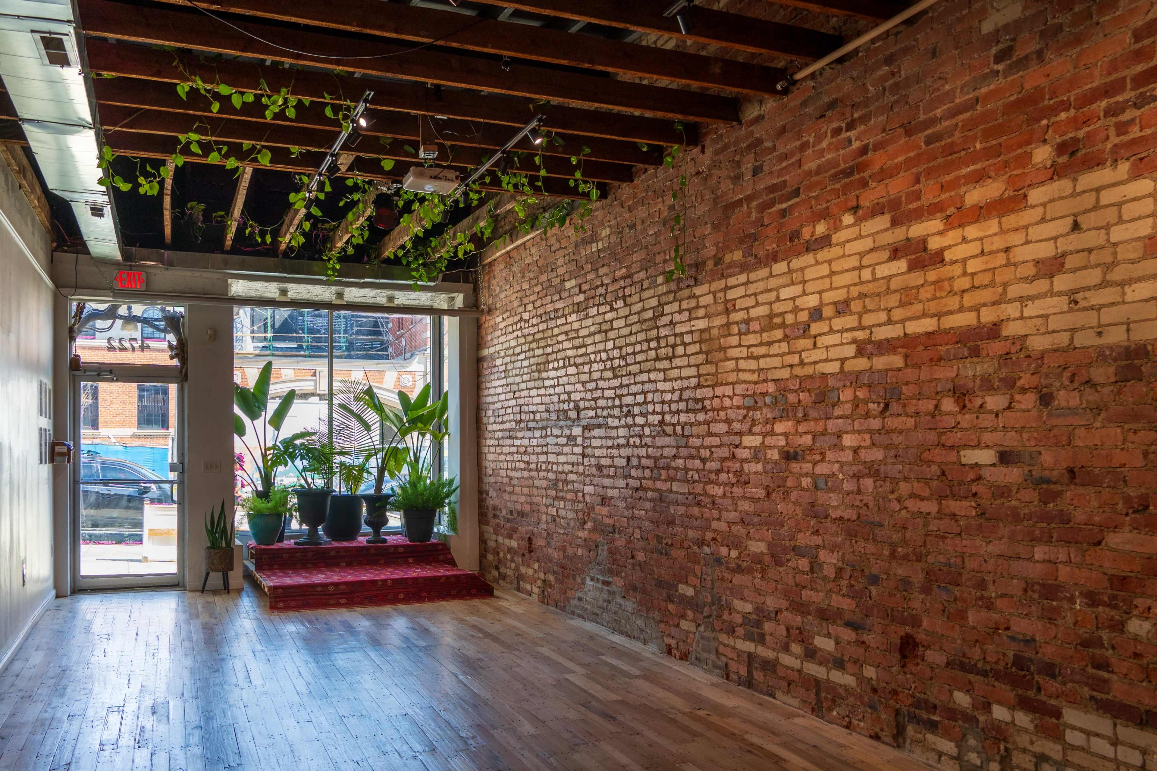 The image shows an empty room with exposed brick walls, wooden flooring, and several potted plants near a doorway that leads outside.