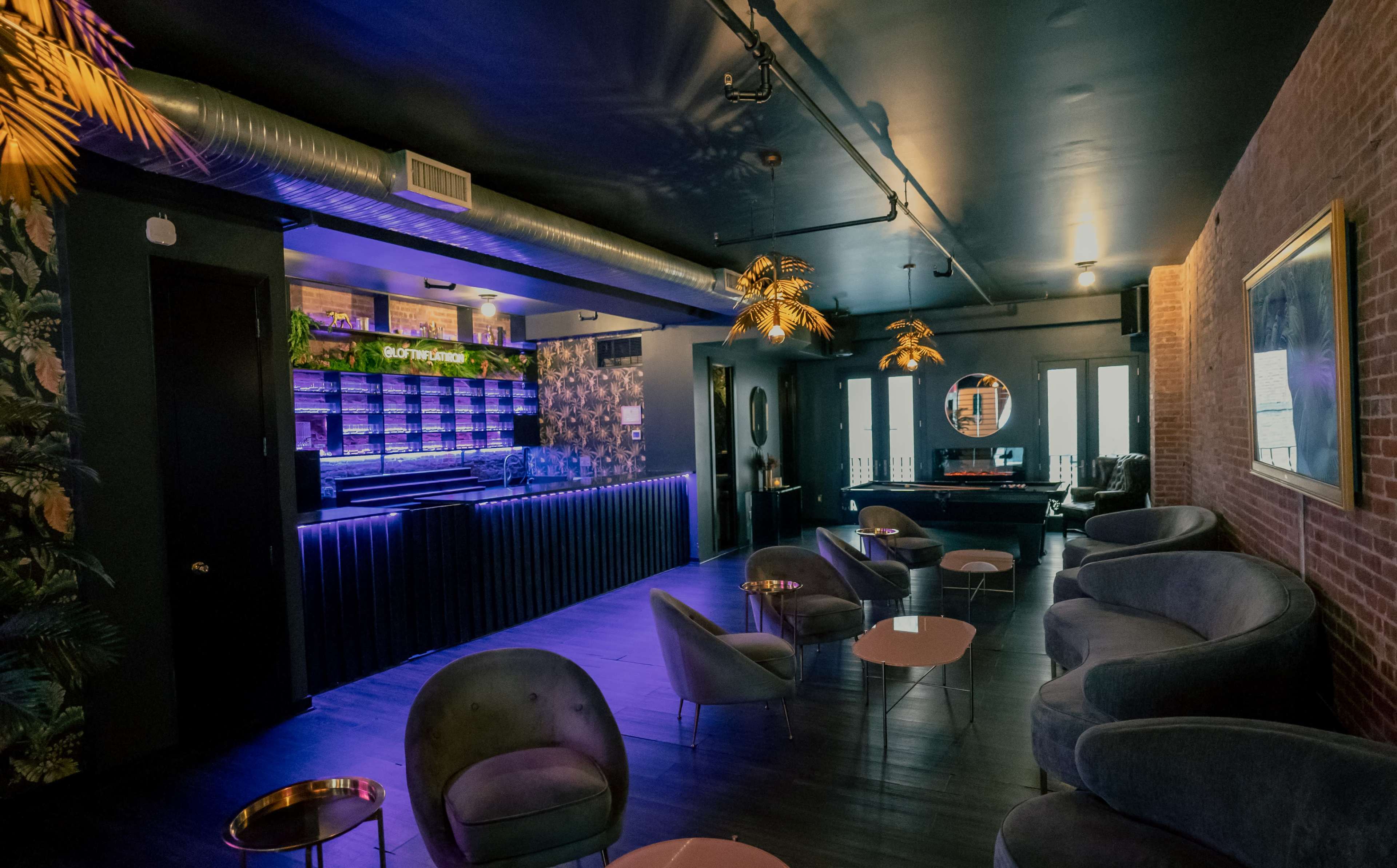 The image shows a modern lounge with plush gray seating, a bar with neon lighting, and decorative plants on the walls.