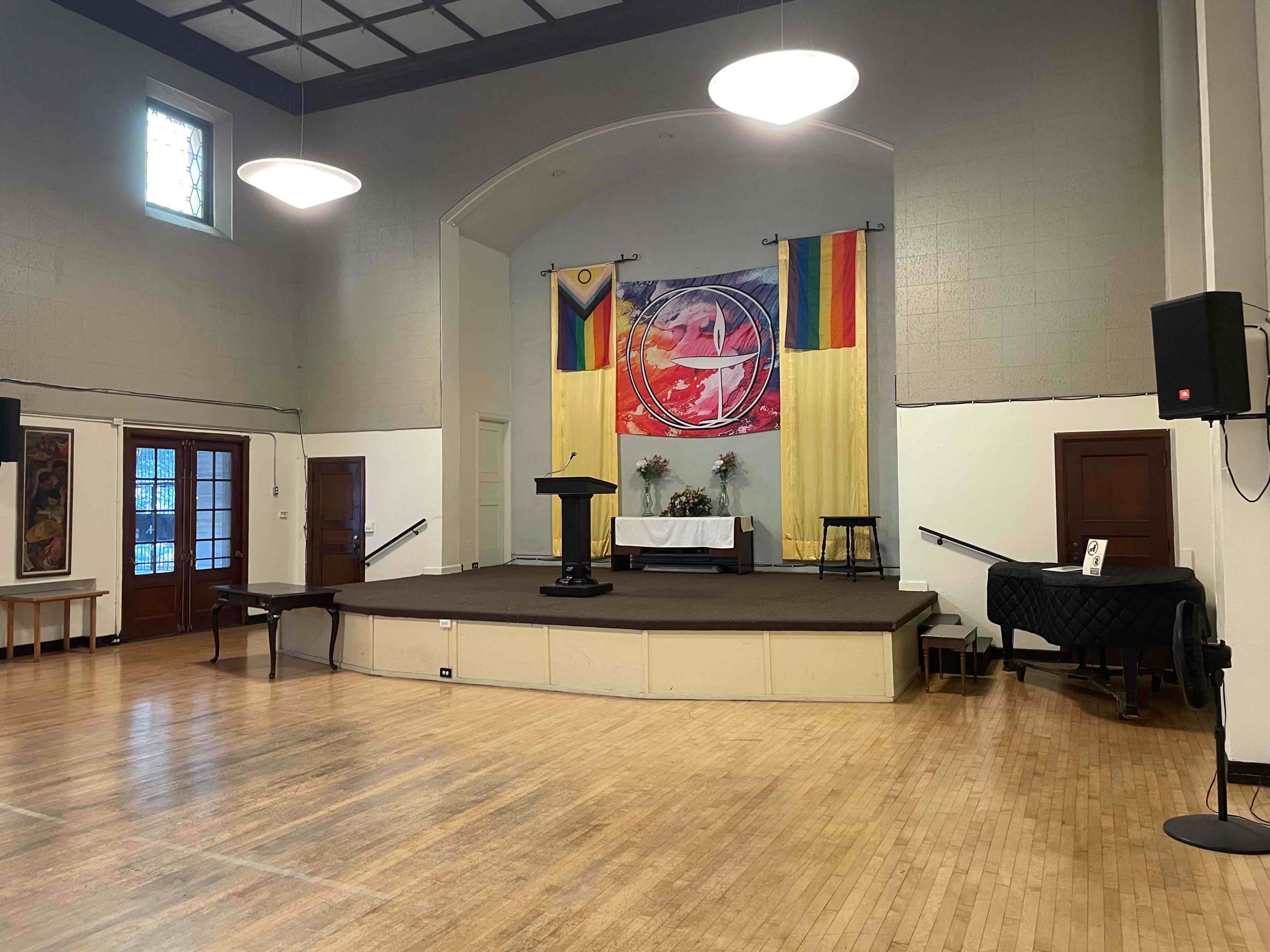 The image shows a spacious interior of a hall featuring a stage with a podium and chairs, decorated with a large banner displaying rainbow colors and other symbols.