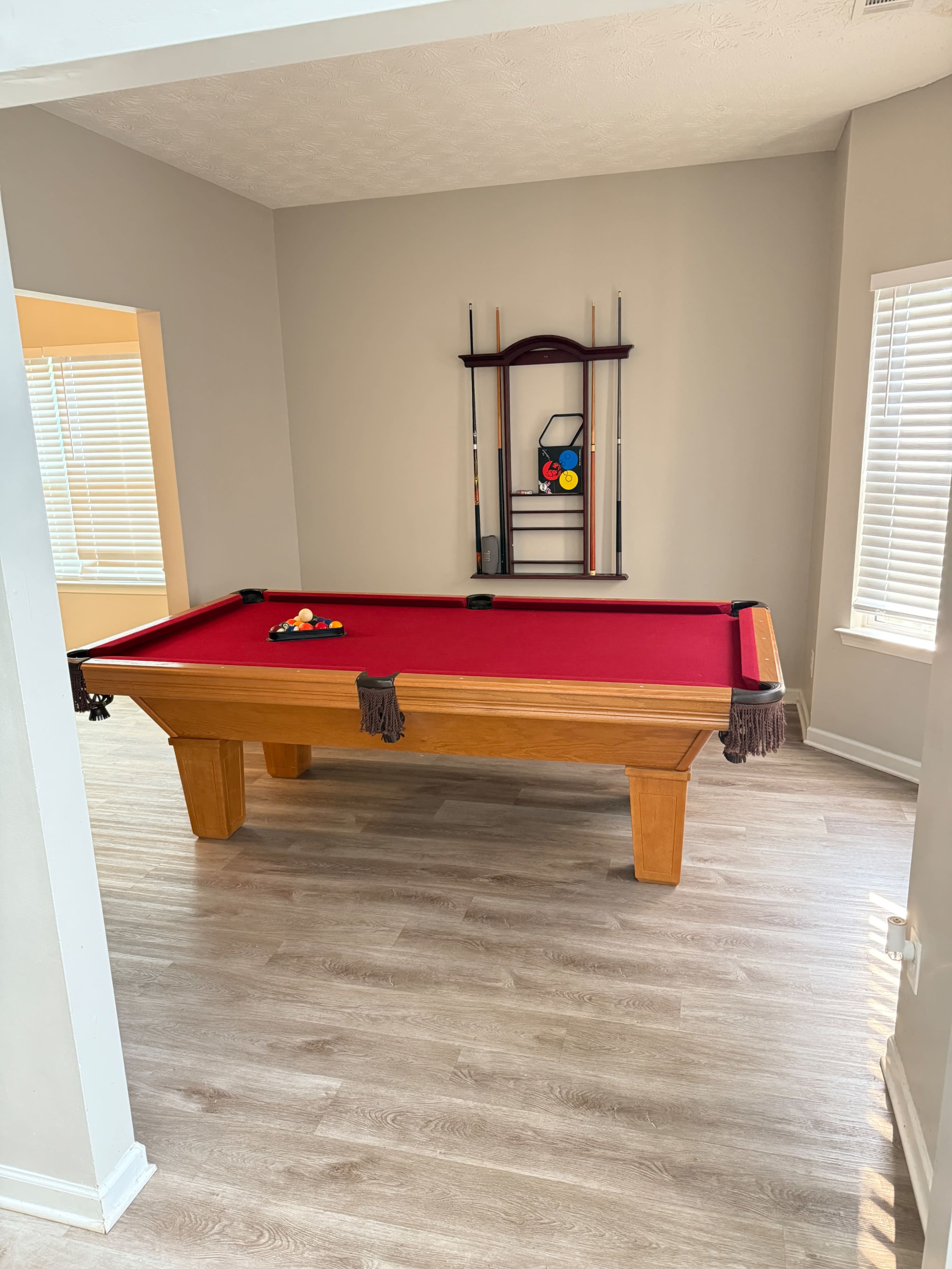 A red-felted billiards table is positioned in a well-lit room with beige walls and wooden floors, accompanied by a wall-mounted rack holding pool cues.