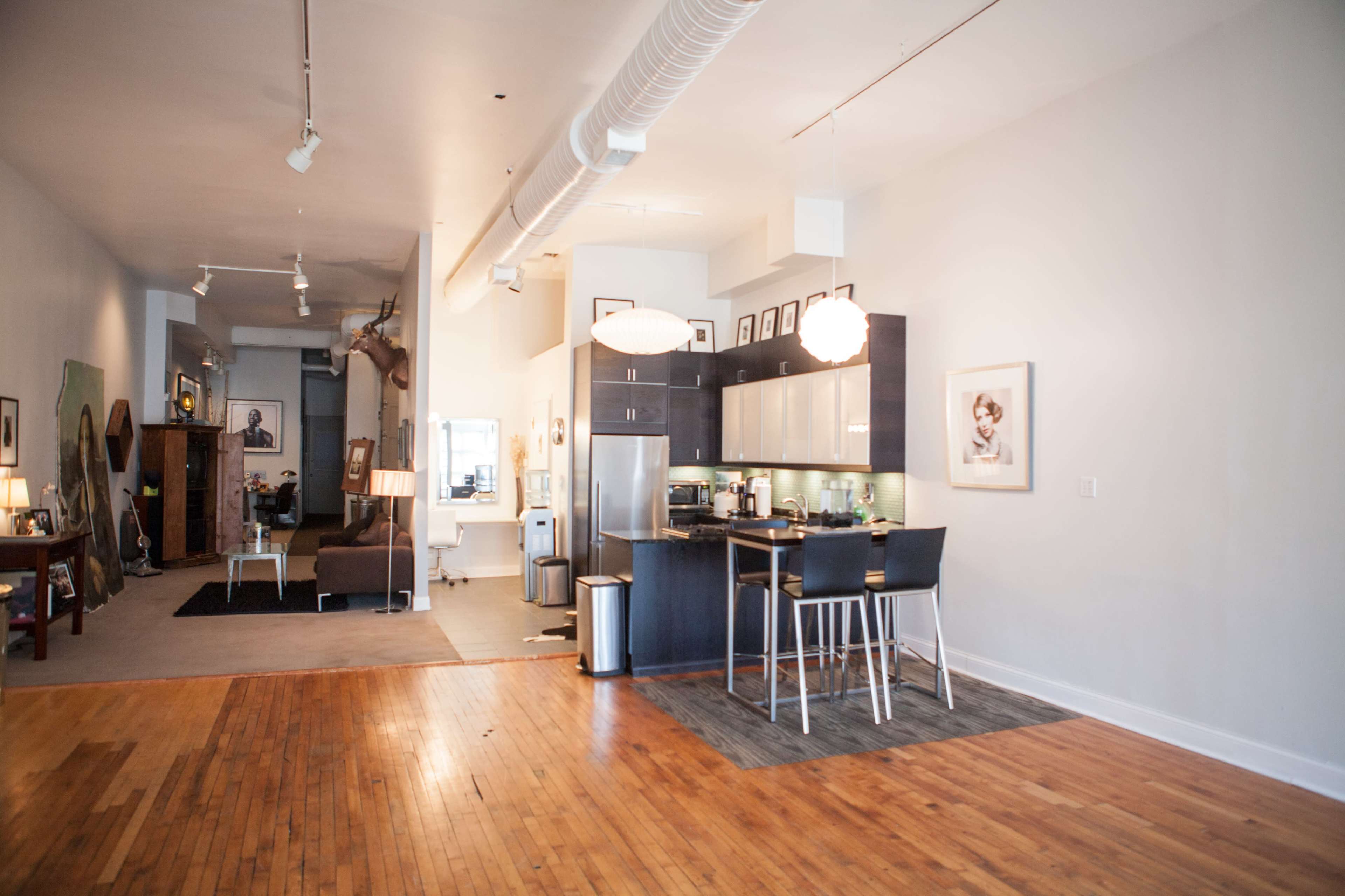 The image shows a spacious open-concept loft featuring a modern kitchen with dark cabinetry and two bar stools, adjacent to a living area with a couch and various decorative items.