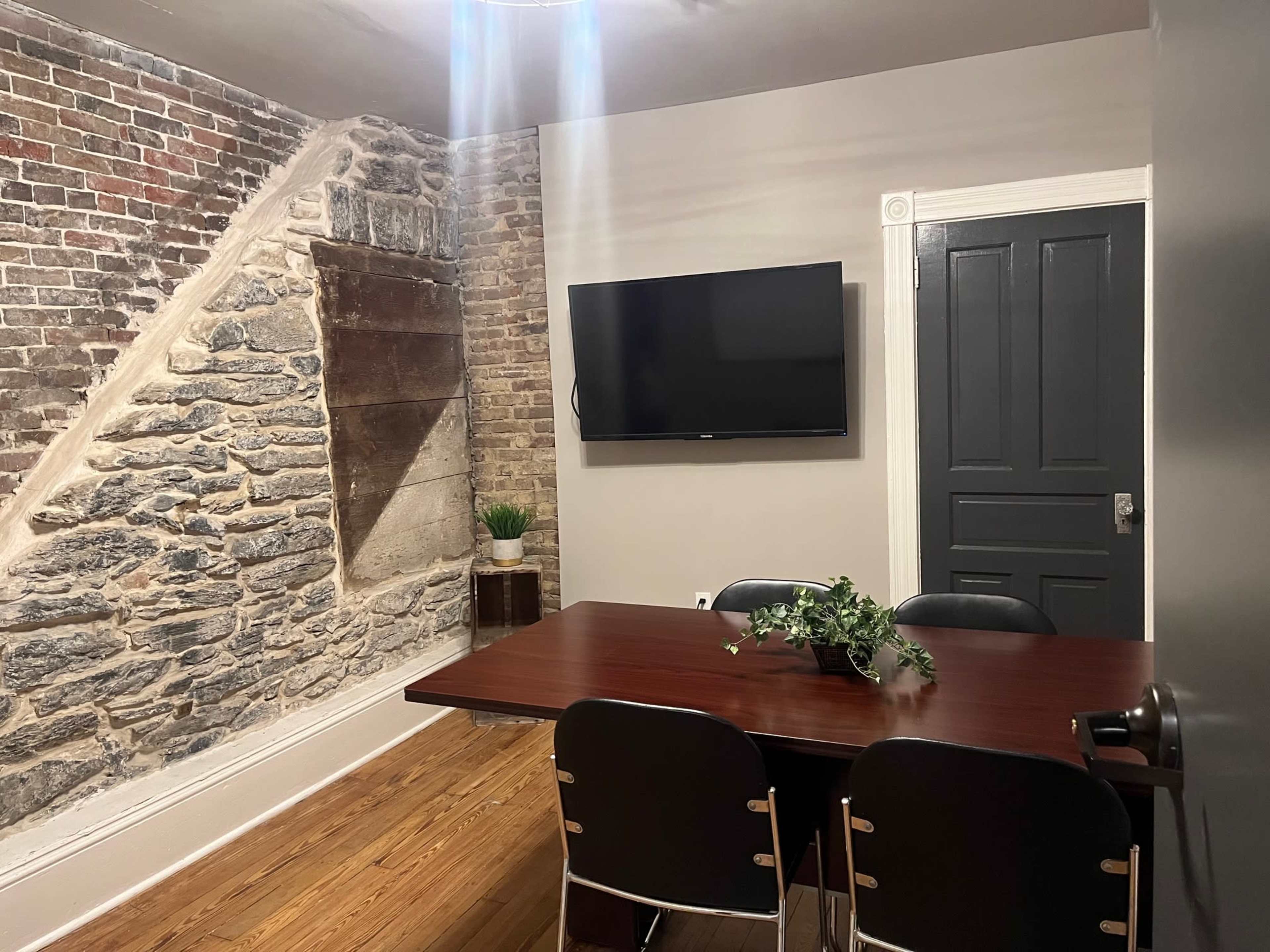 The image shows a dining area featuring a table with several black chairs, a flat-screen TV mounted on the wall, and a combination of exposed brick and stone walls.