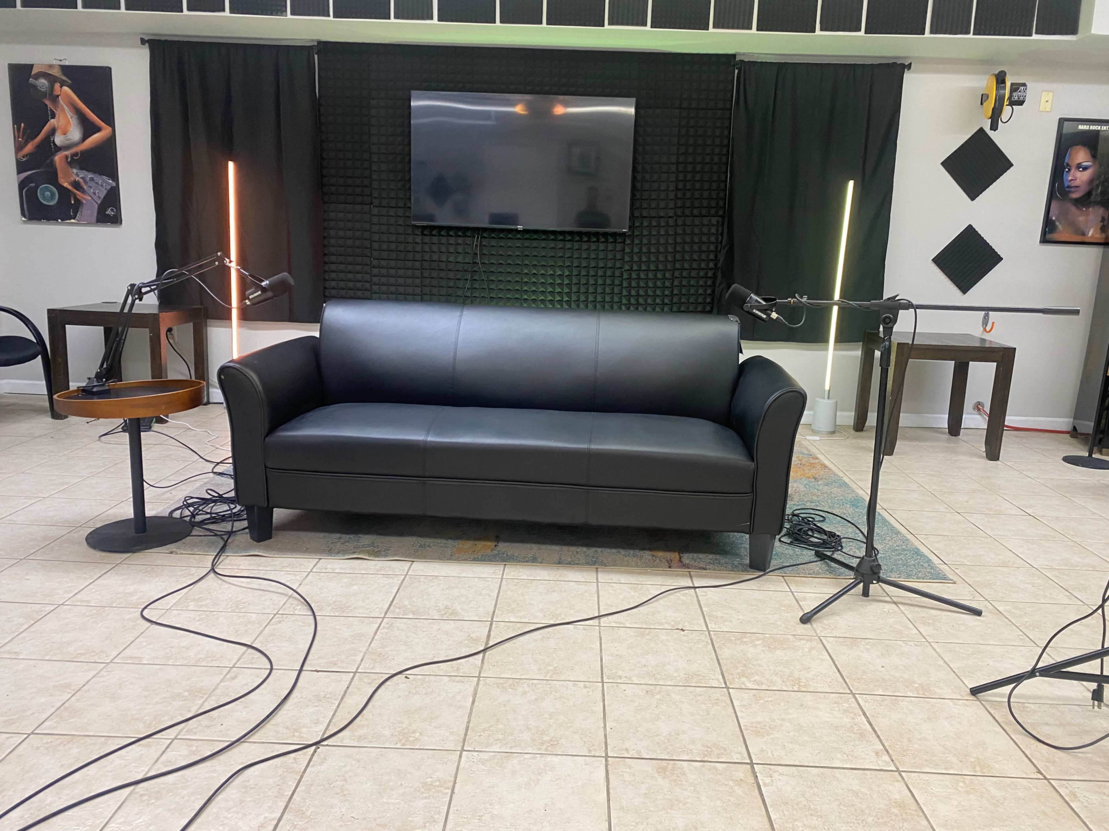 A modern living space with a black couch in the center, flanked by two standing lights, surrounded by microphones on stands and artwork on the walls.
