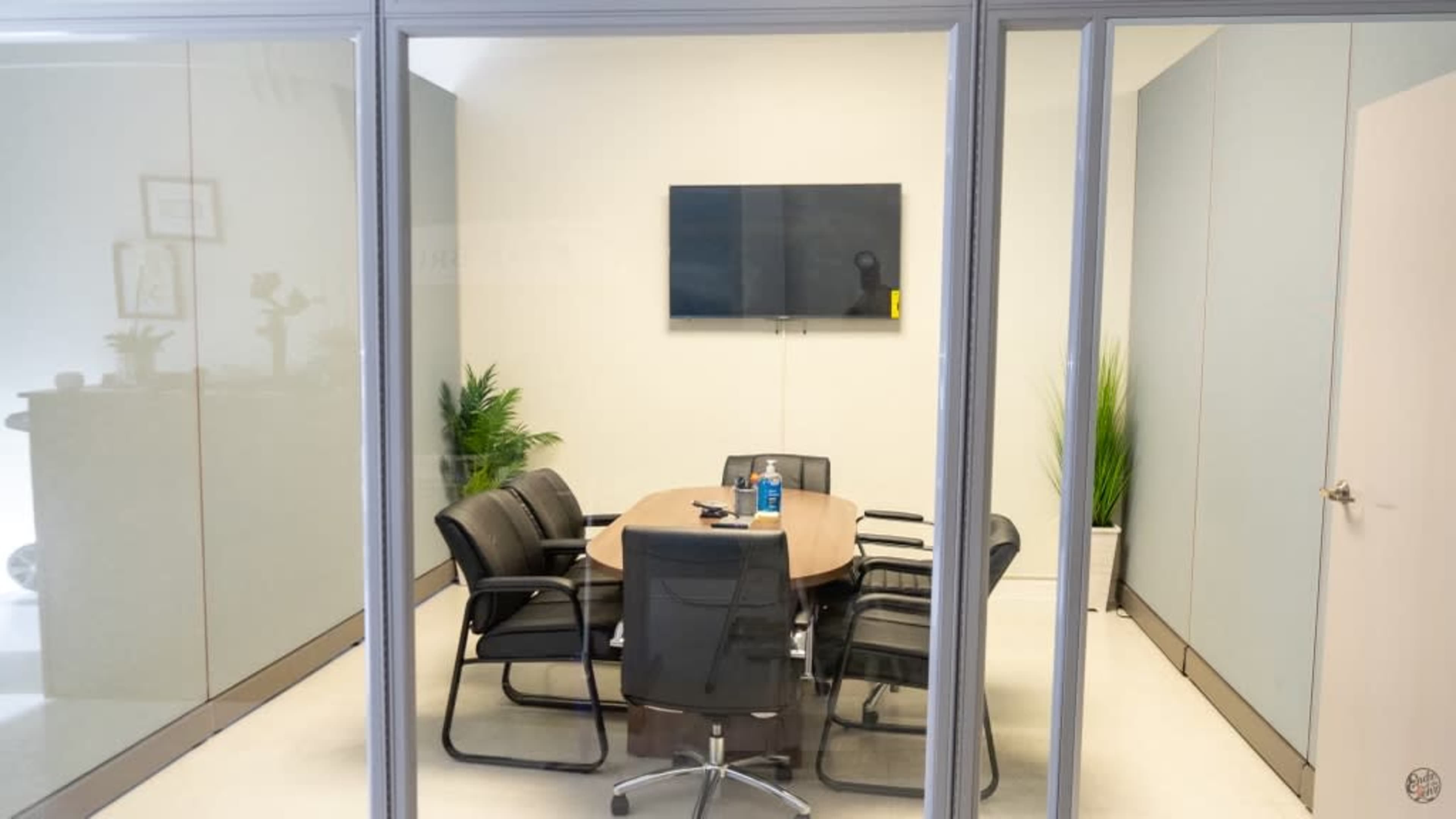 A conference room is visible through glass walls, featuring a round table surrounded by six chairs and a wall-mounted television.