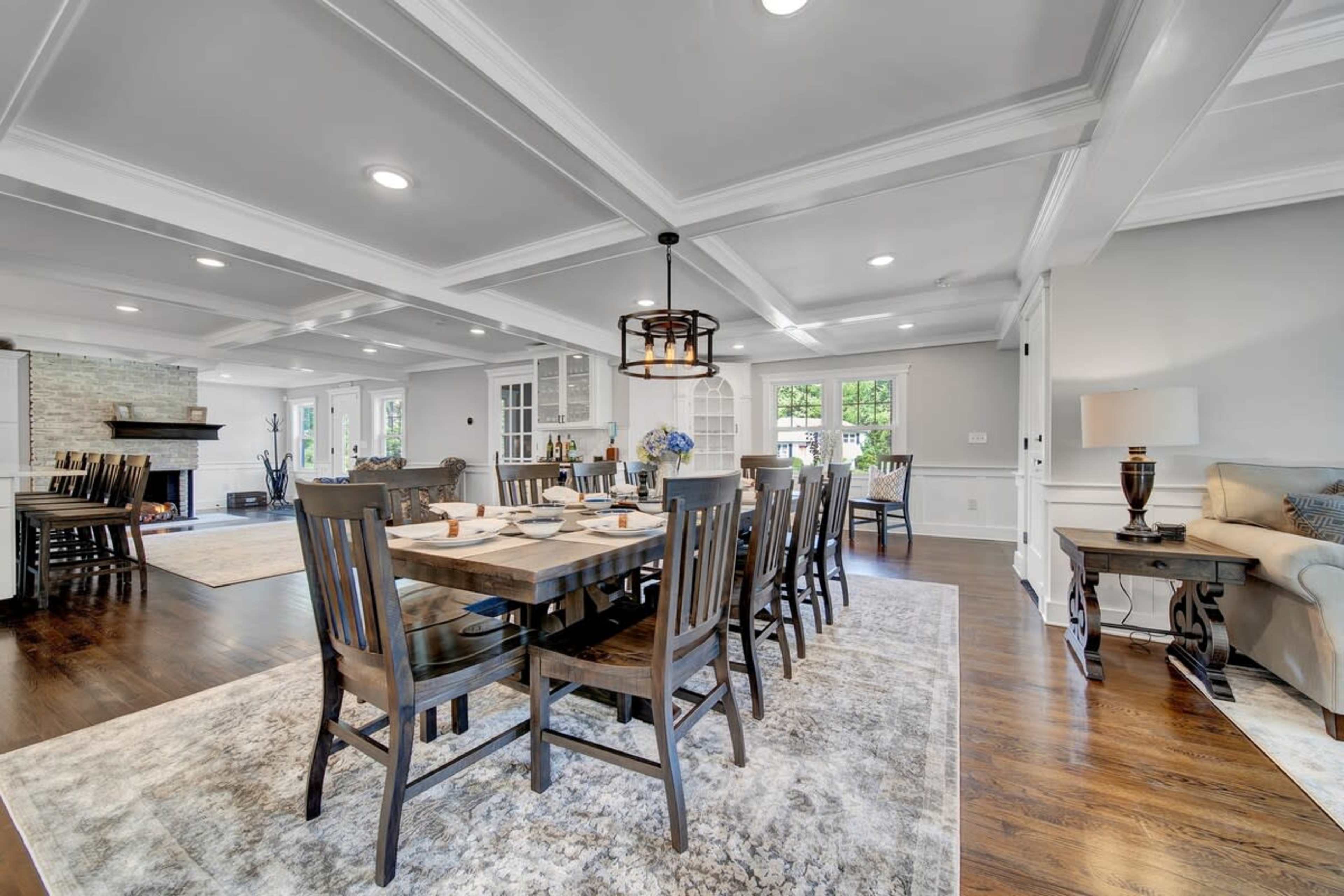 A spacious dining area with a large wooden table surrounded by dark chairs, adjacent to a living room with a sofa and a rug.