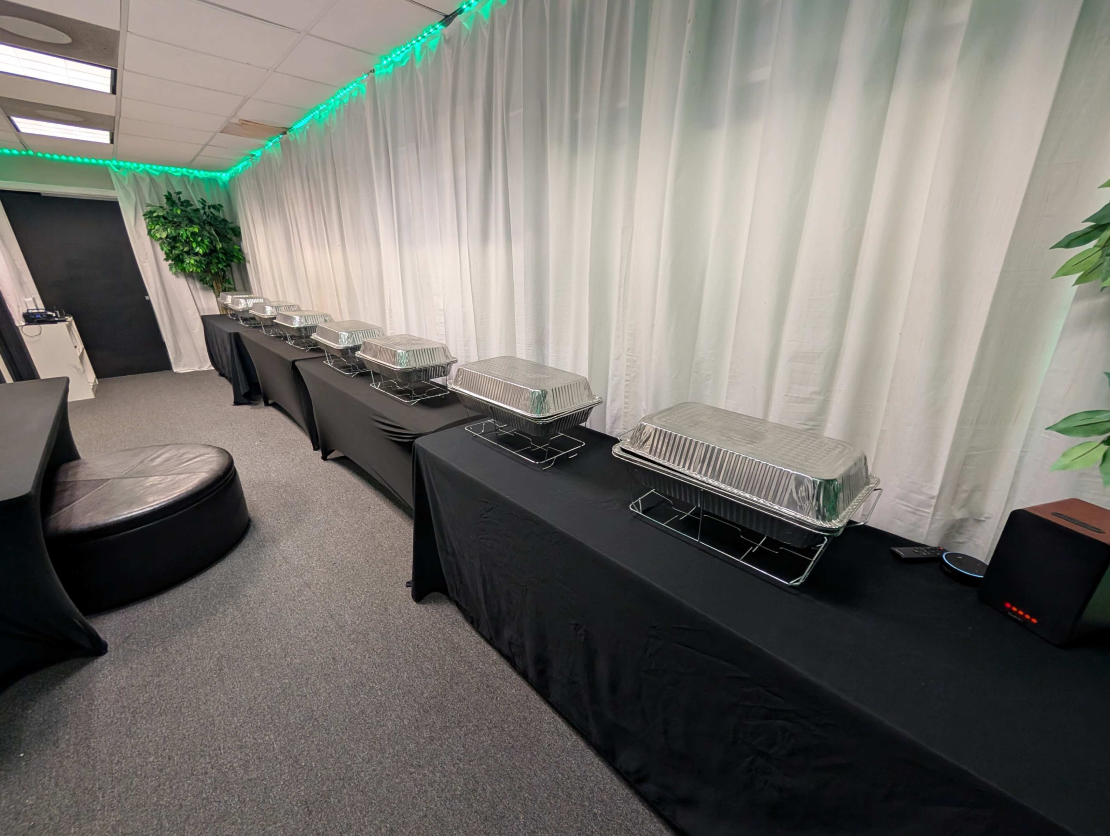 The image shows a line of covered food trays set on black tables beneath green ambient lighting in a room with white curtains.