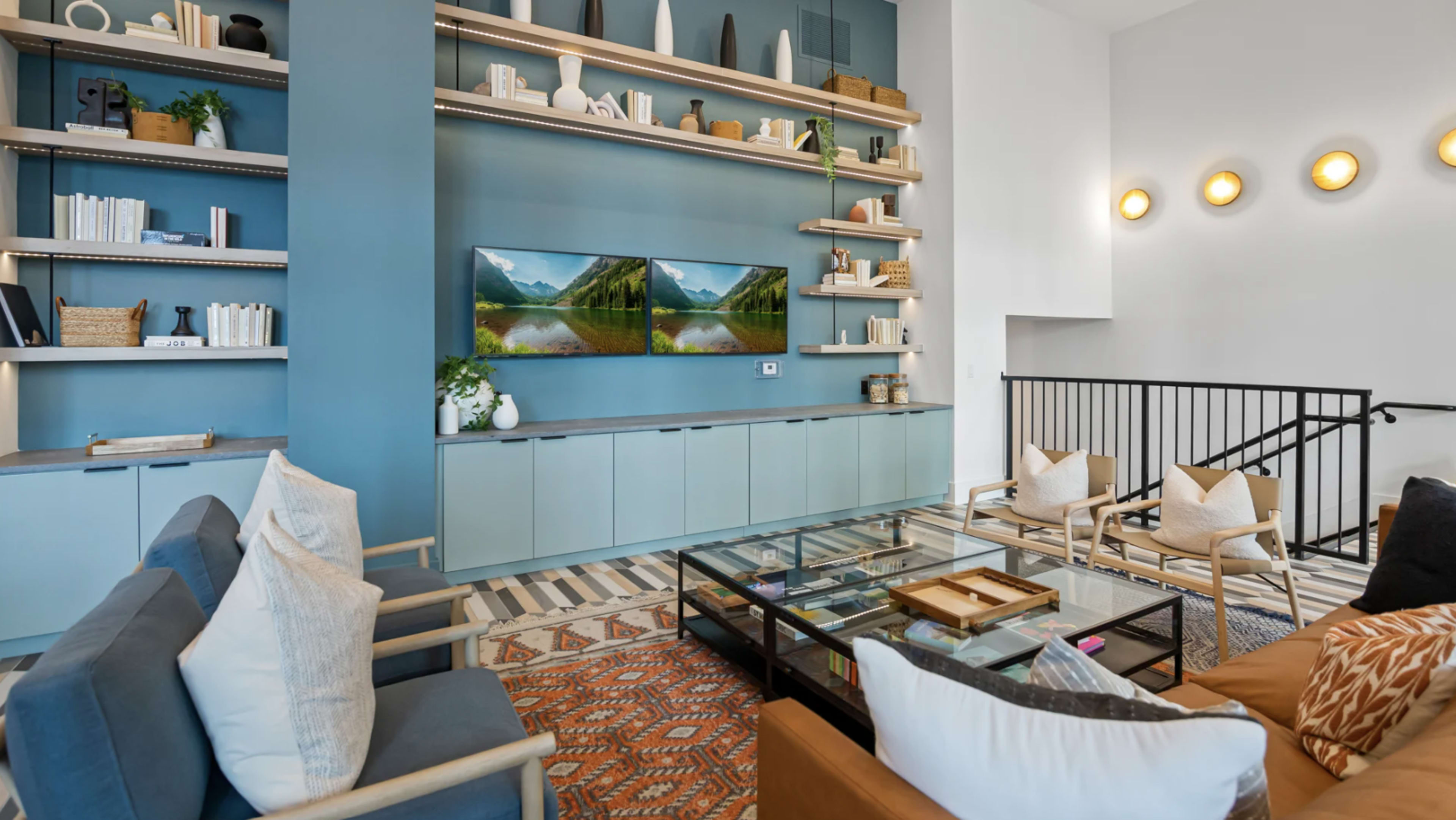 A modern living room features shelves with decorative items, a large glass coffee table, and two wall-mounted televisions above a light blue cabinet.