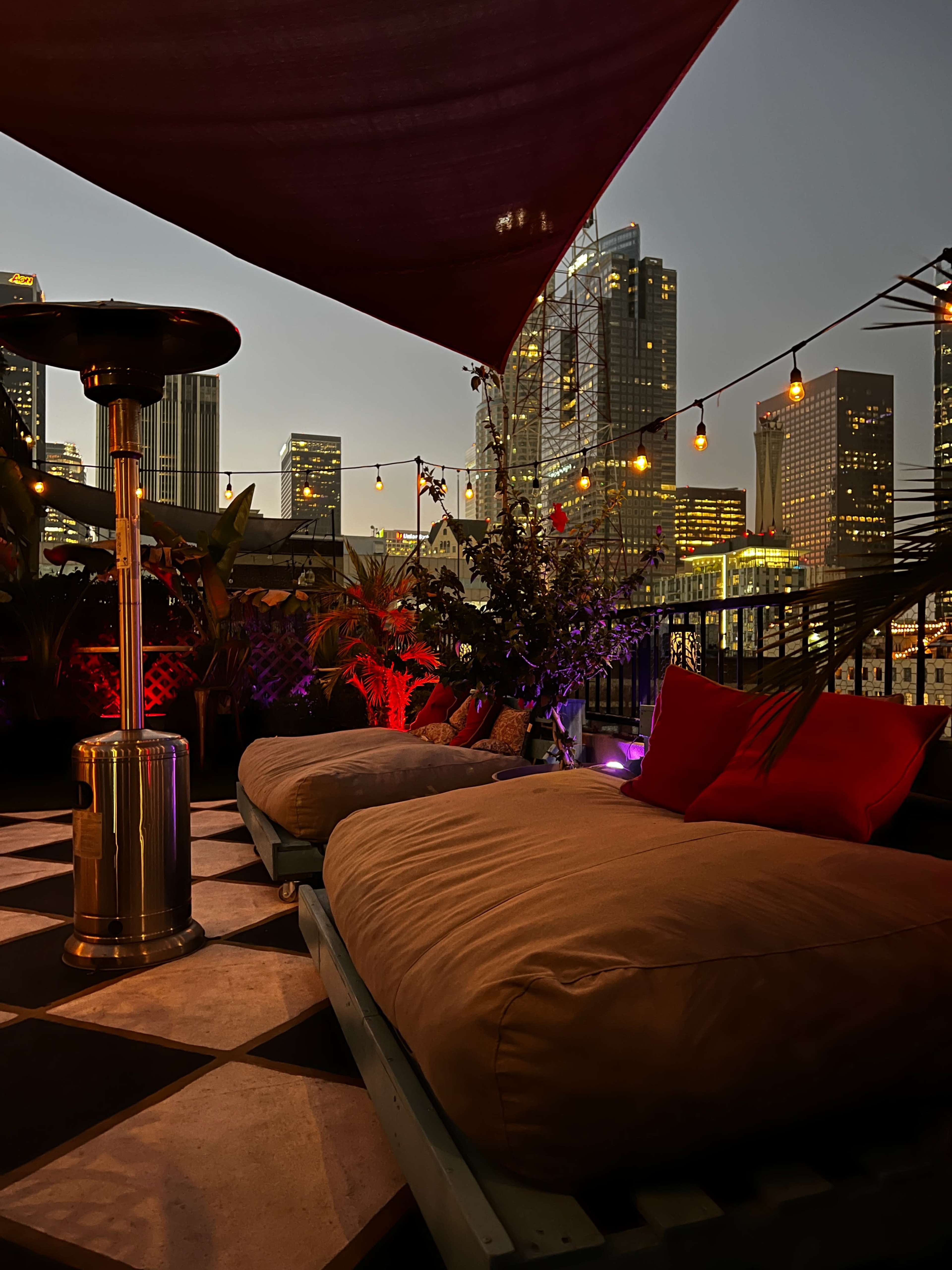 A rooftop seating area features oversized cushions, a heater, and string lights, with city skyscrapers visible in the background at dusk.