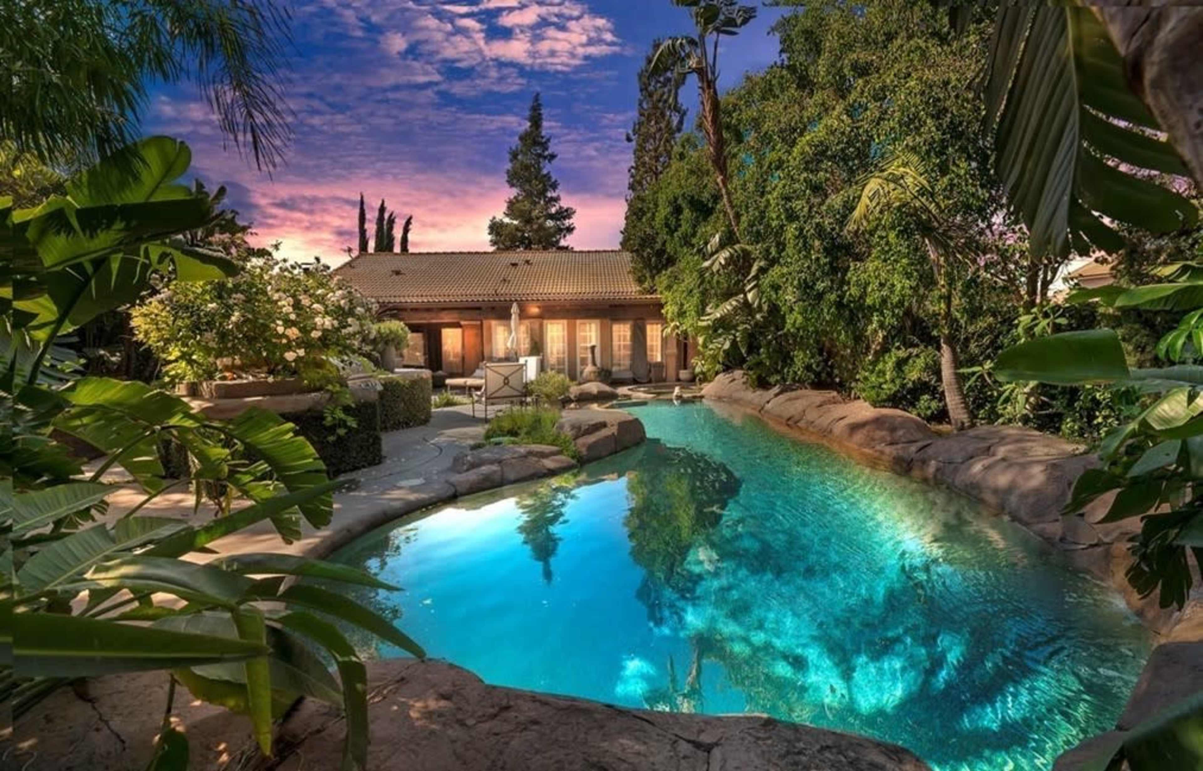 A resort-style swimming pool surrounded by lush greenery and a backdrop of a house at sunset.