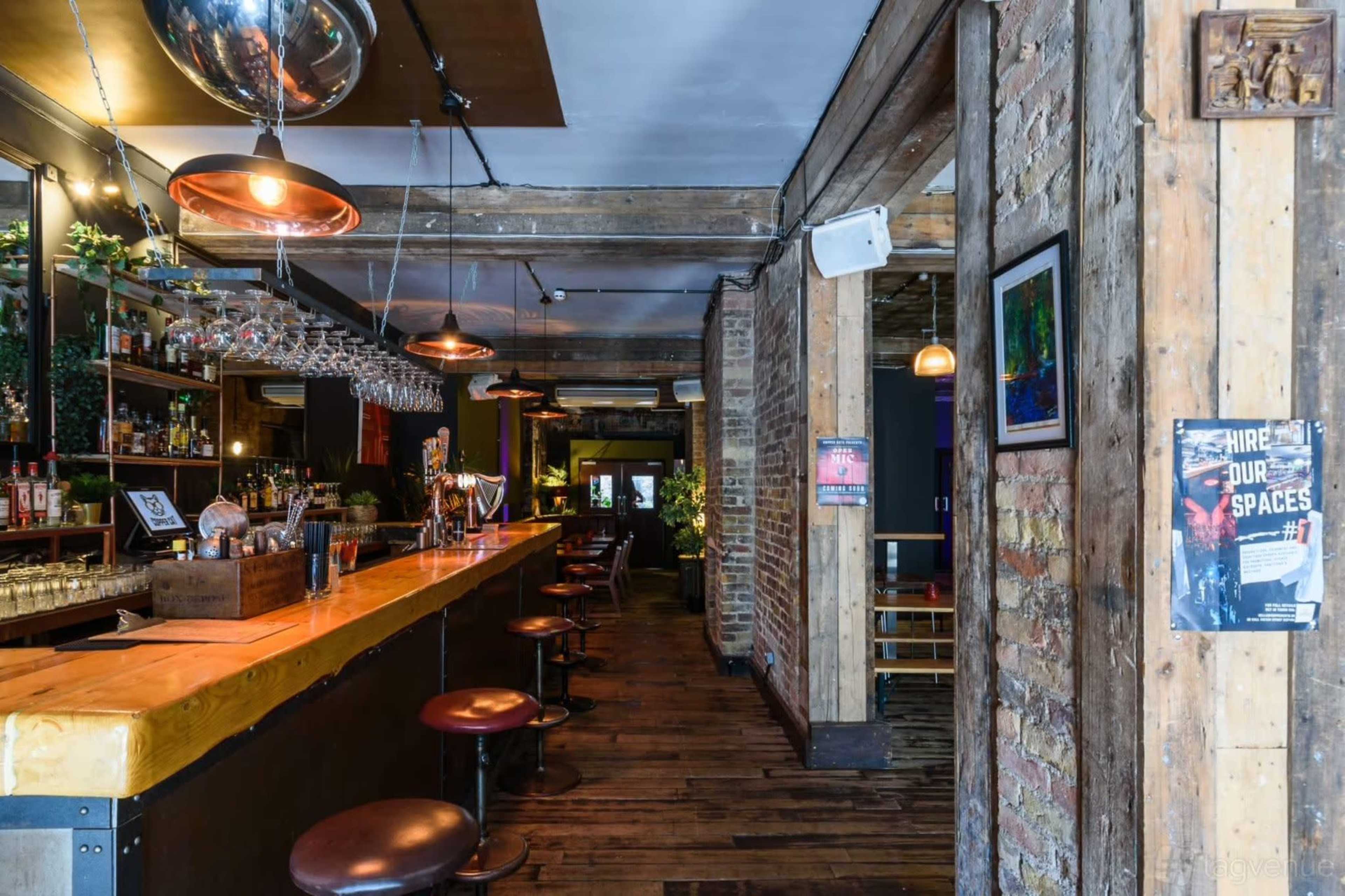 The image shows the interior of a bar with wooden beams, a long counter, and tall chairs, illuminated by pendant lights.