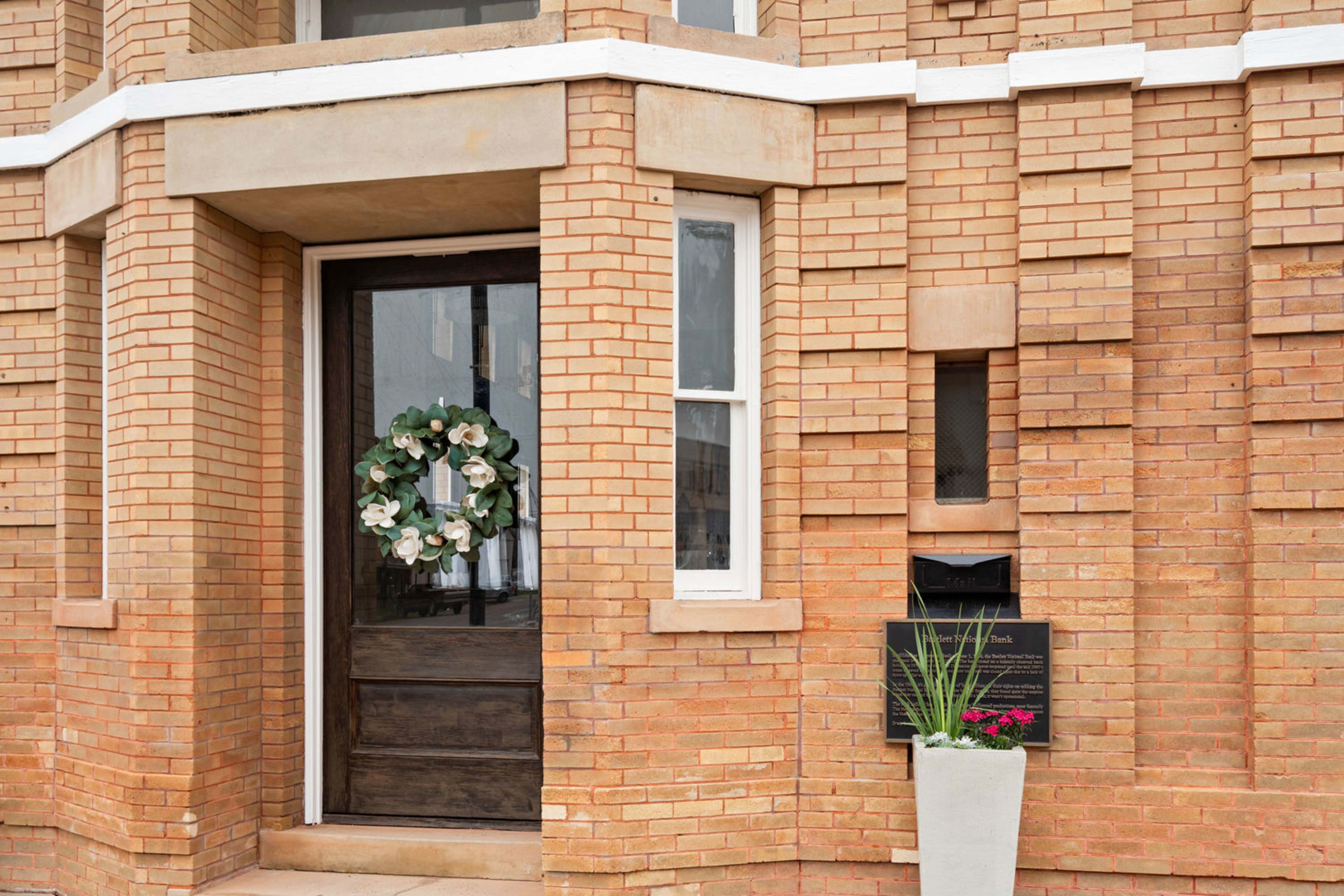 A brick building entrance with a wooden door, decorated with a wreath, and a planter with flowers beside a mailbox.
