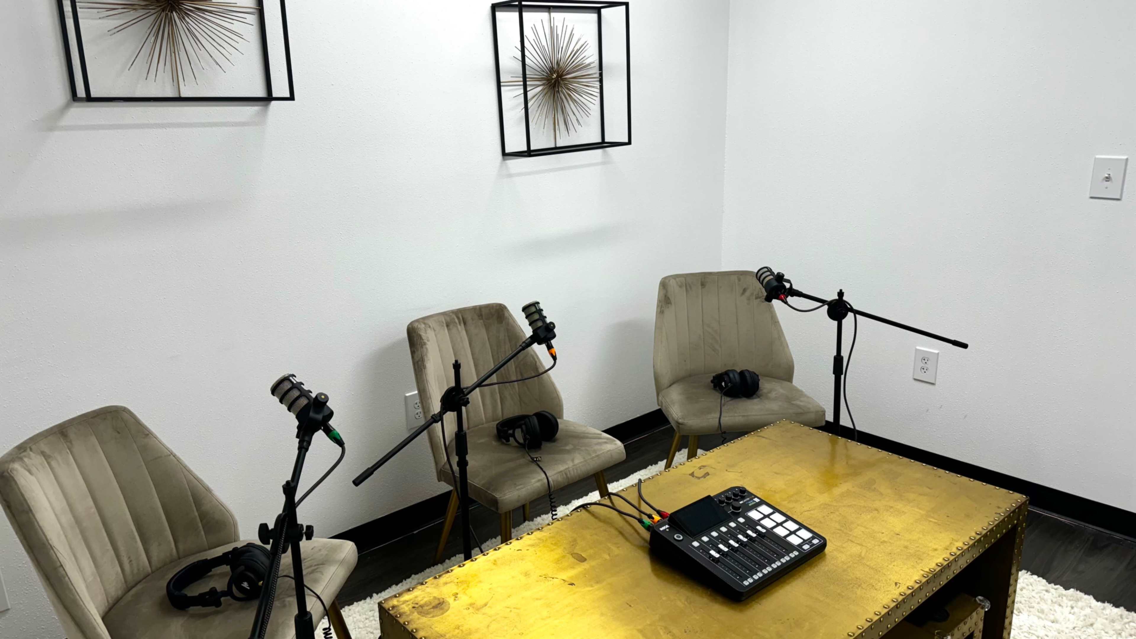 The image shows a podcasting studio with three chairs, each equipped with a microphone and headphones, and a sound mixing console on a gold table.