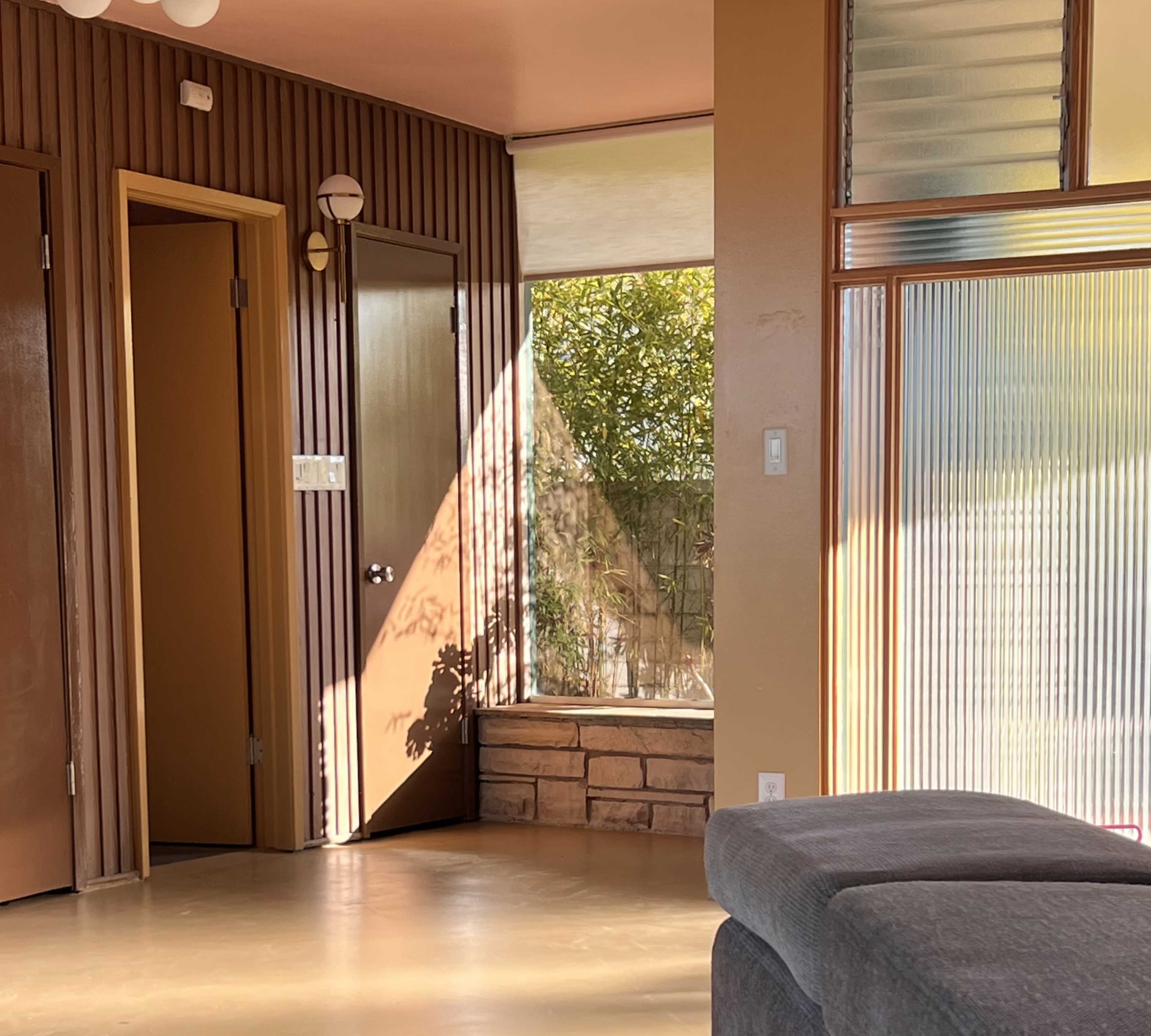 The image shows a sunlit interior space with brown paneling, a doorway, and a window revealing green foliage outside.
