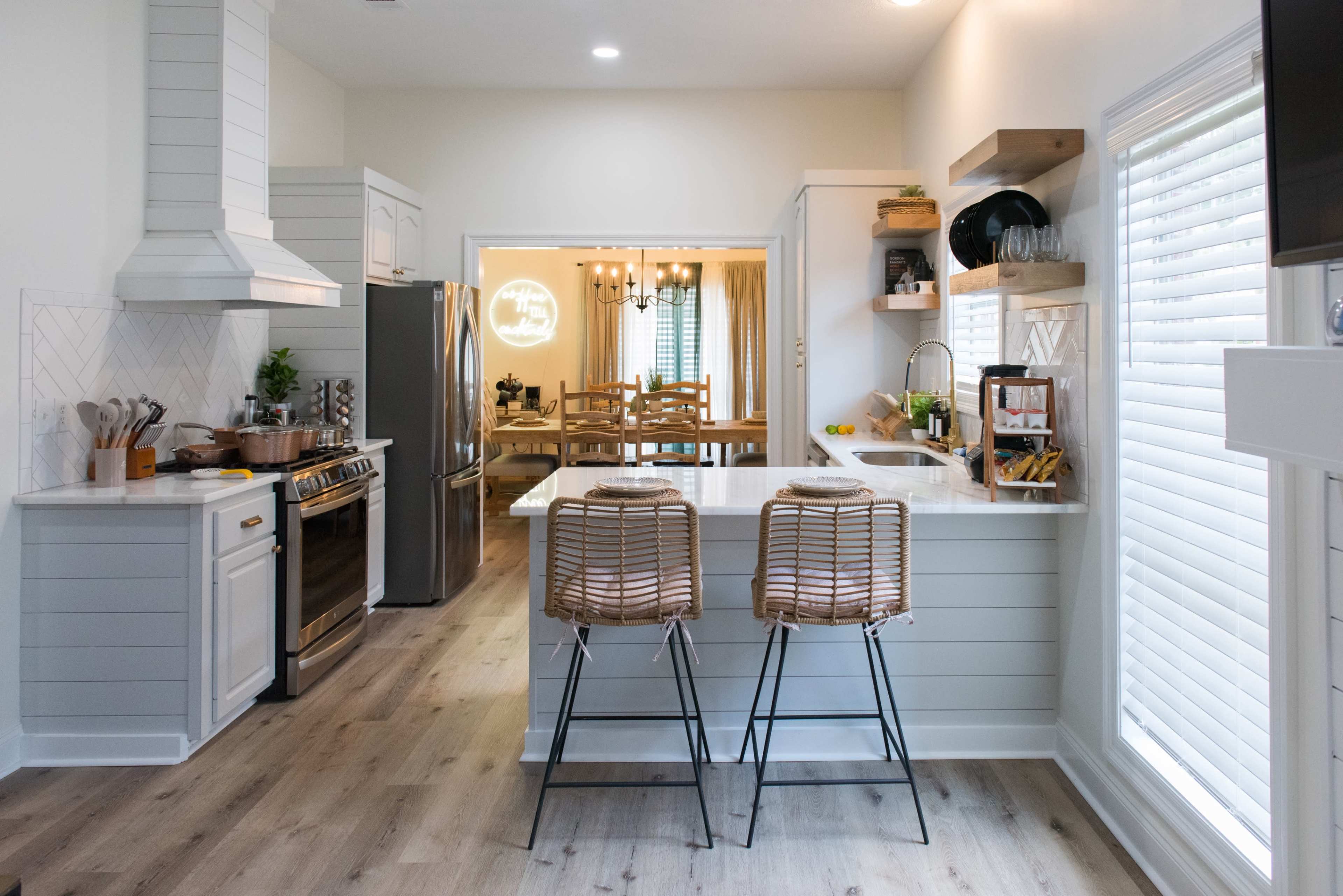 A modern kitchen with light-colored cabinetry, stainless steel appliances, and two barstools at a kitchen island, leading into a dining area with wooden furniture.