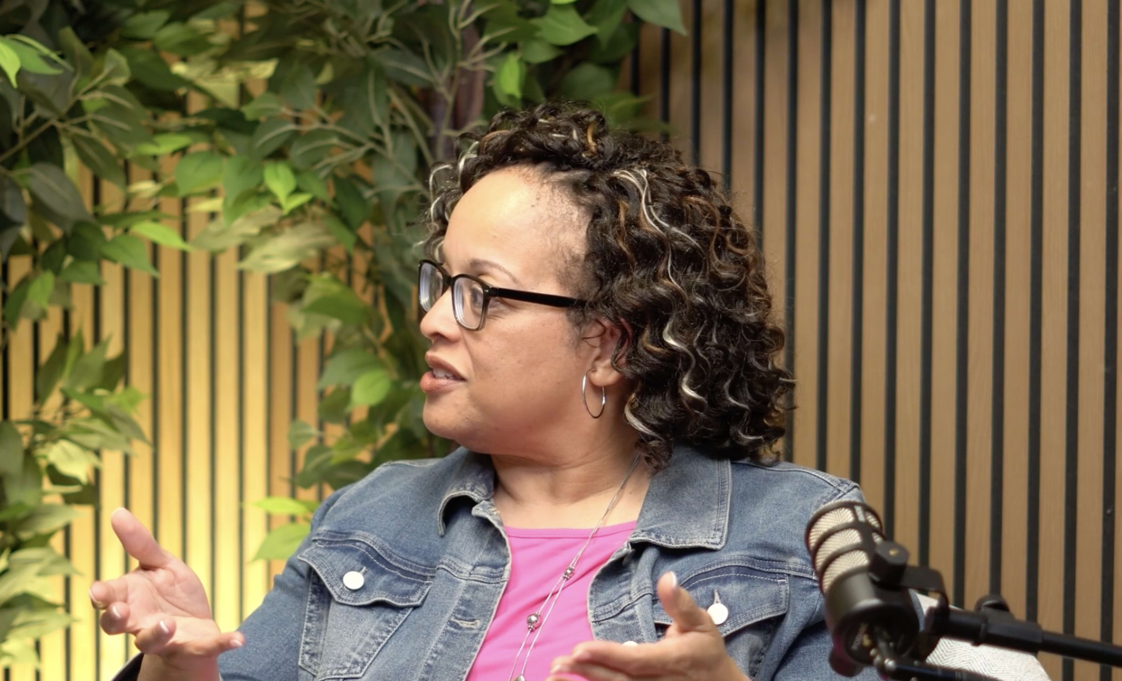 A woman with curly hair and glasses is speaking while seated in front of a backdrop of greenery and wooden paneling.