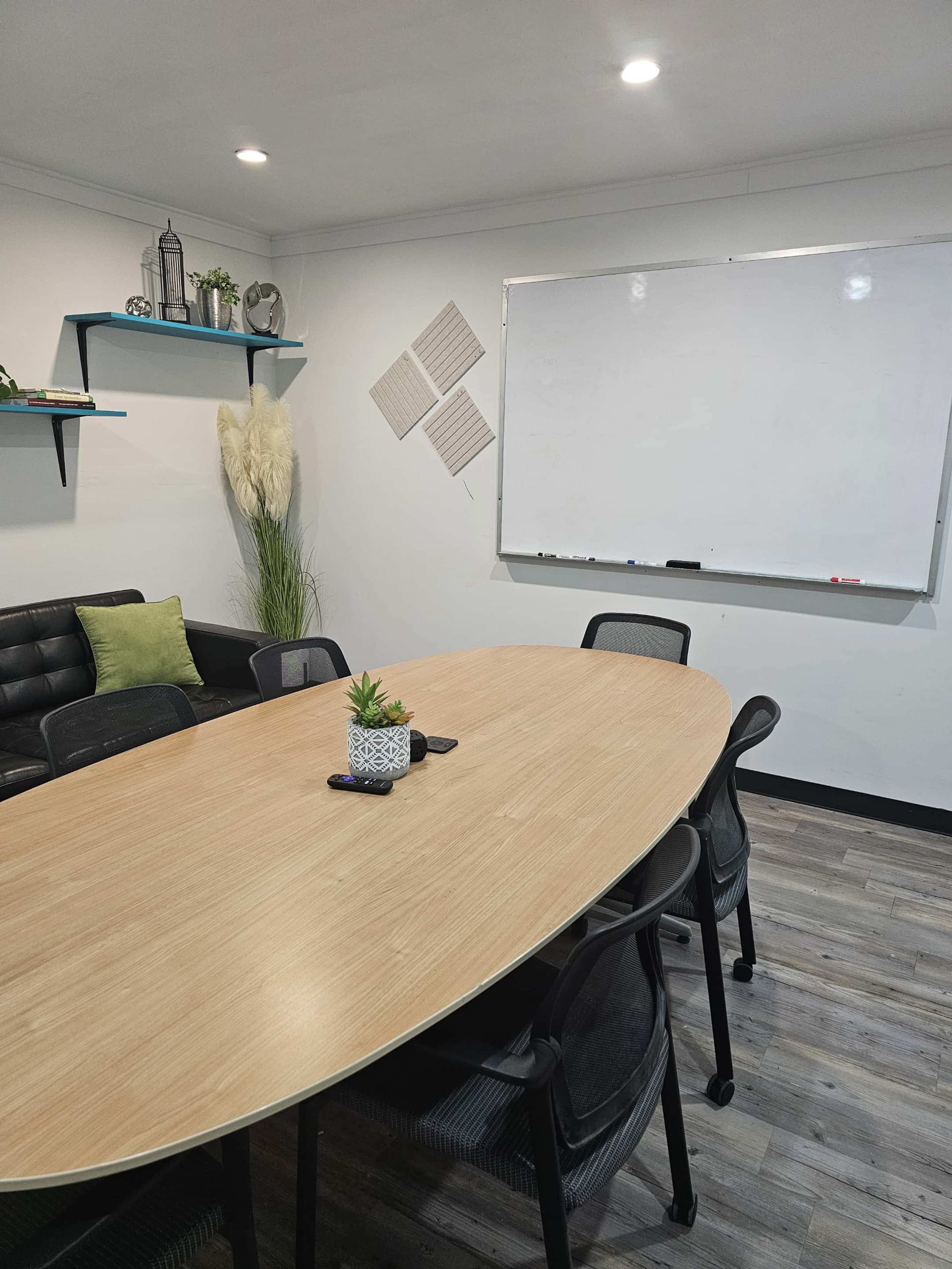 The image shows a modern conference room with a large oval wooden table surrounded by black chairs, a whiteboard on the wall, and decorative plants and shelves.