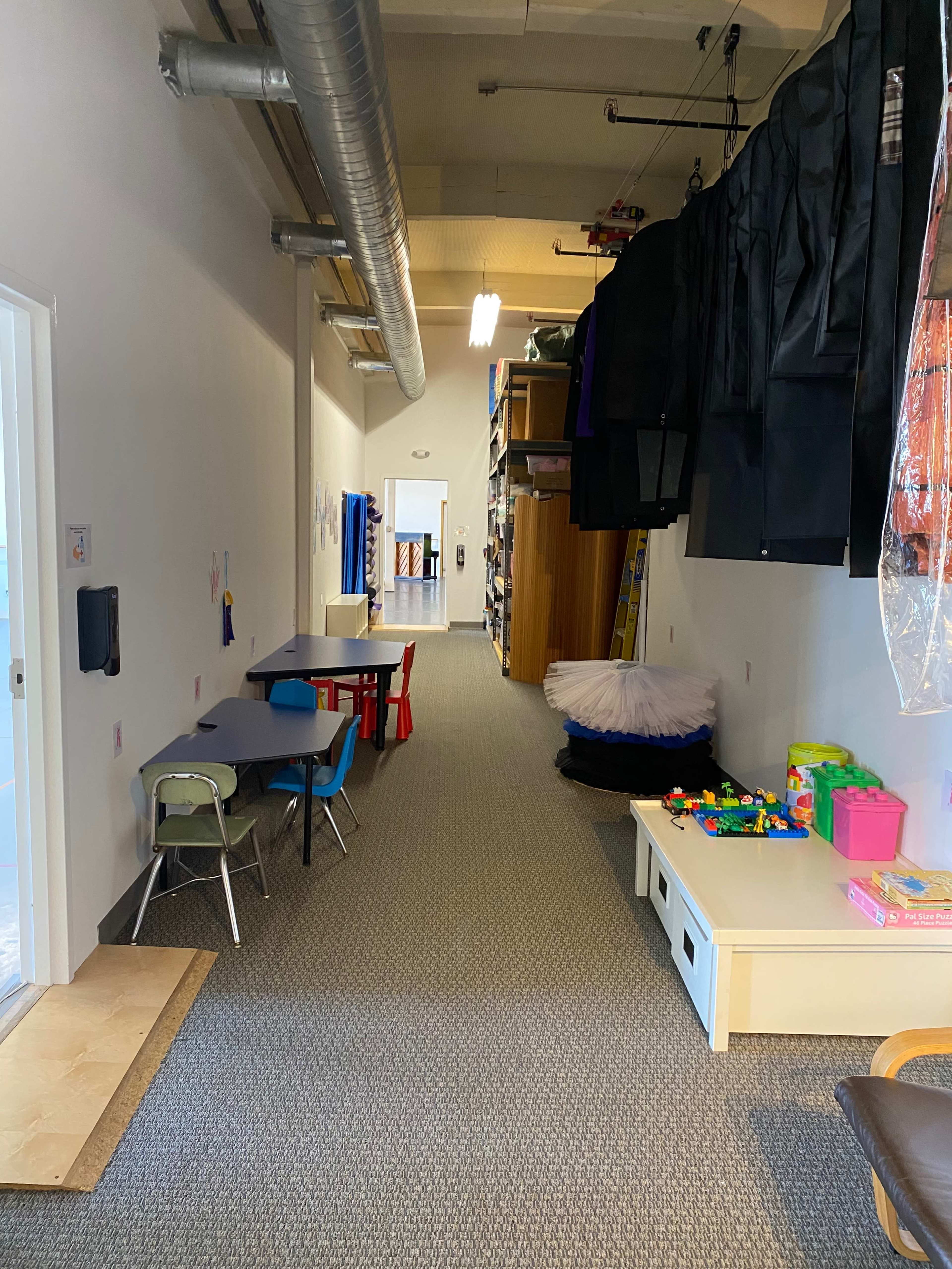 The image shows a long, narrow room with a carpeted floor, lined with storage shelves, a table and chairs on one side, and various toys on a low table.