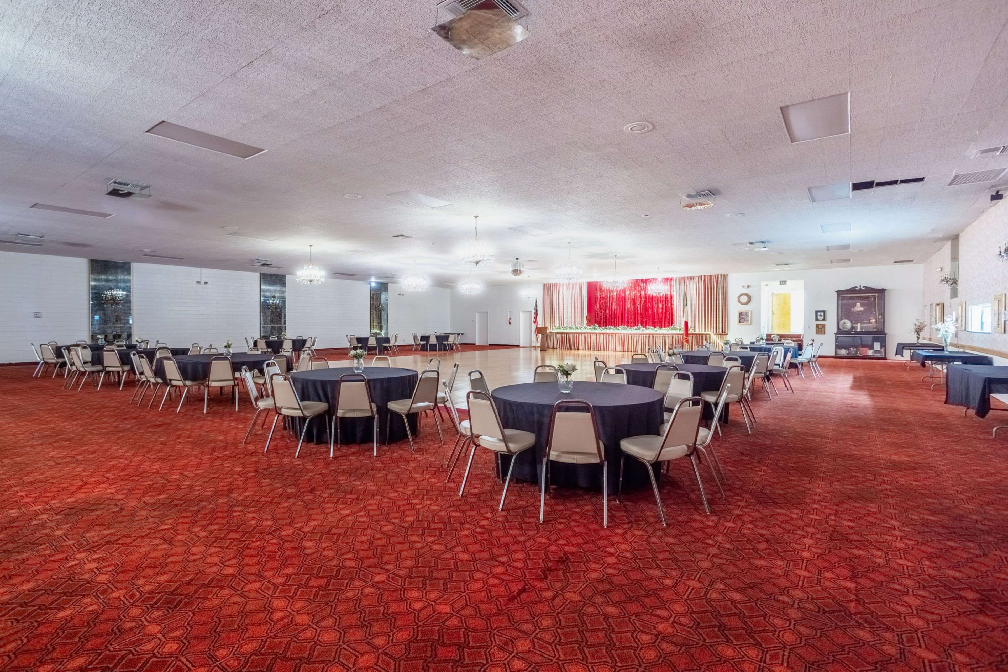 The image shows a spacious banquet hall with round tables covered in dark cloth, arranged on a red carpeted floor, and a stage at the front draped in red curtains.