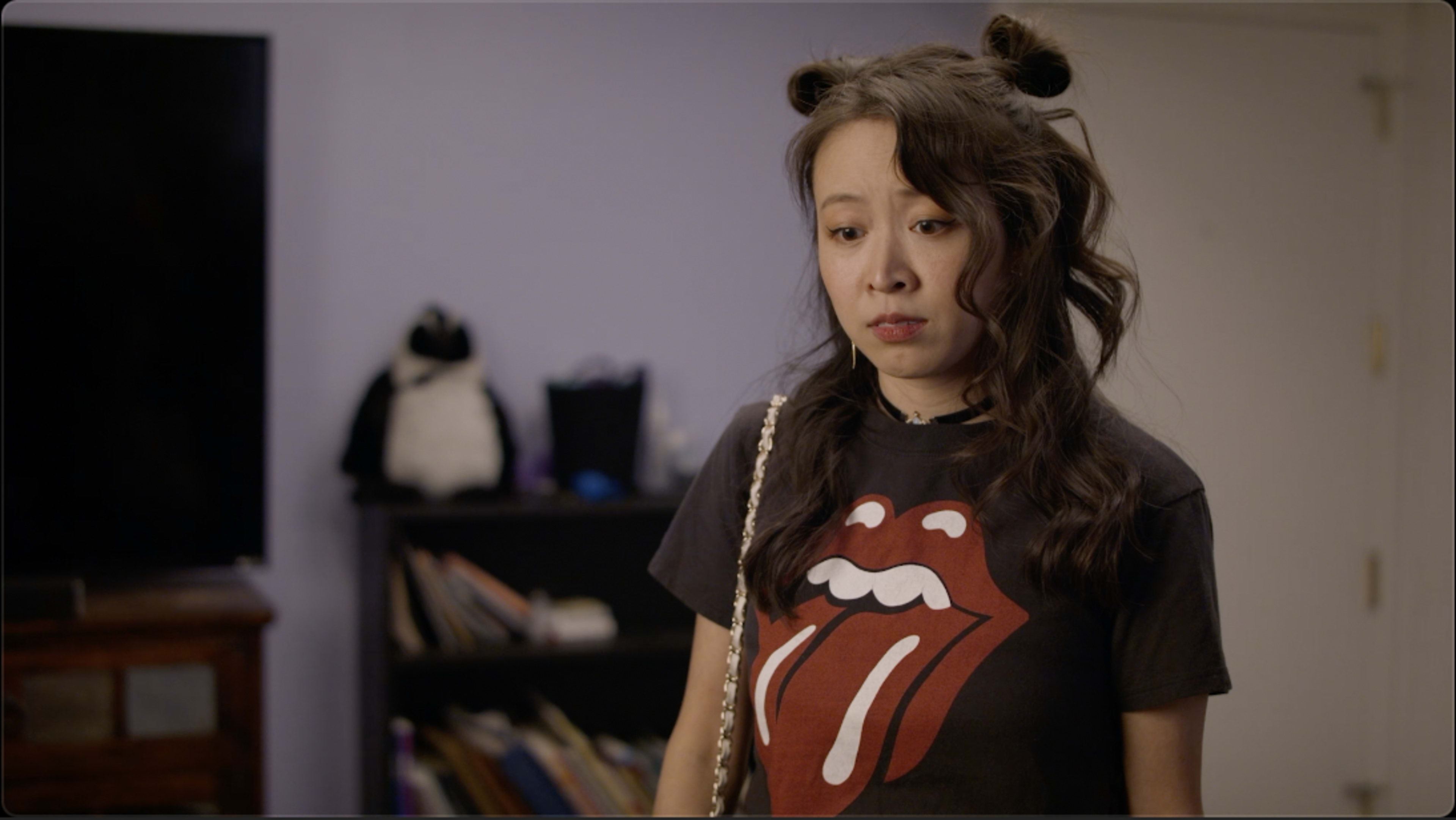 A woman wearing a Rolling Stones t-shirt stands in a room with a television and a bookshelf.