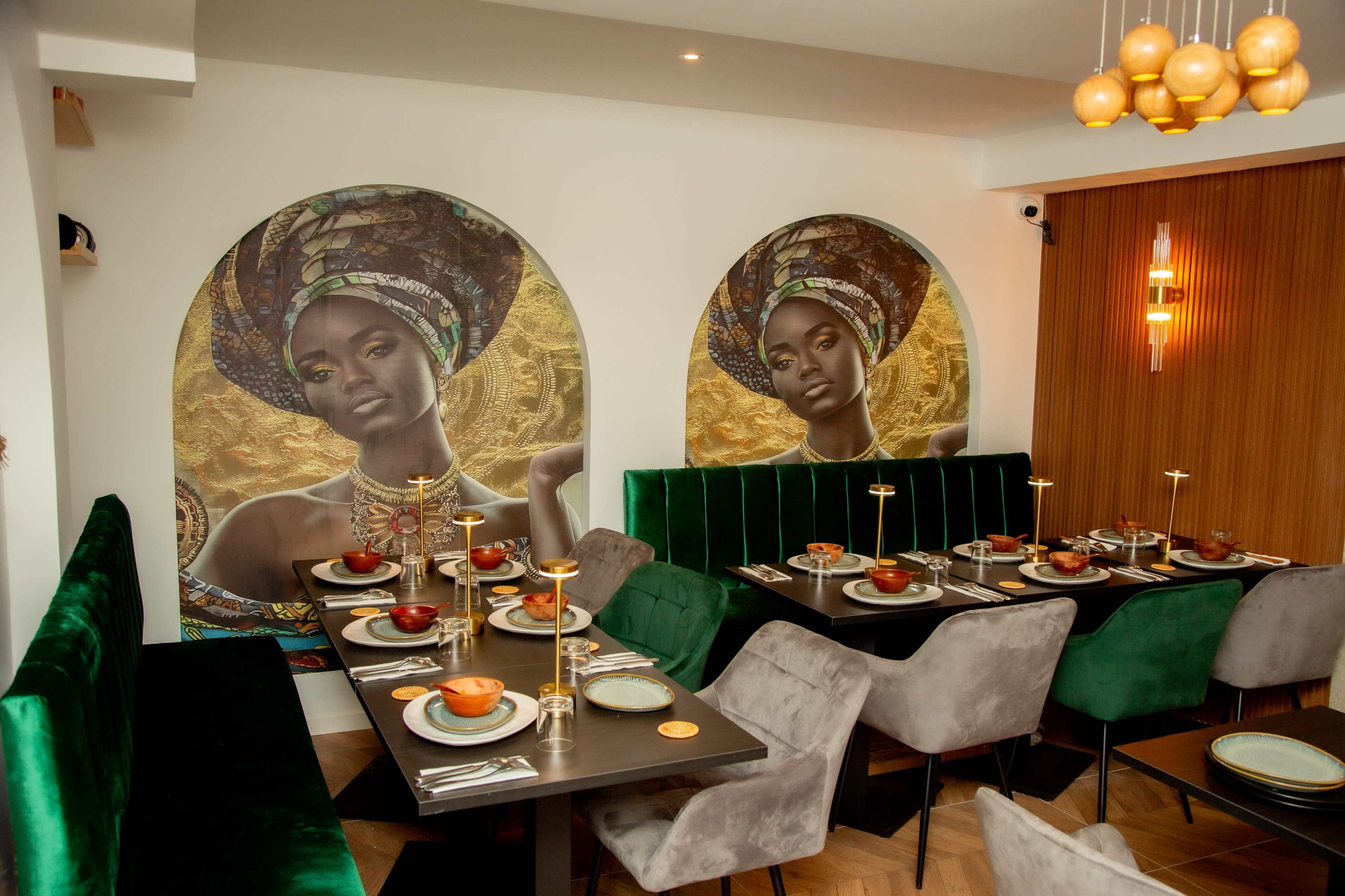The image shows a modern restaurant interior featuring two large murals of women with headwraps, alongside elegantly arranged tables set for dining with dark green and gray chairs.