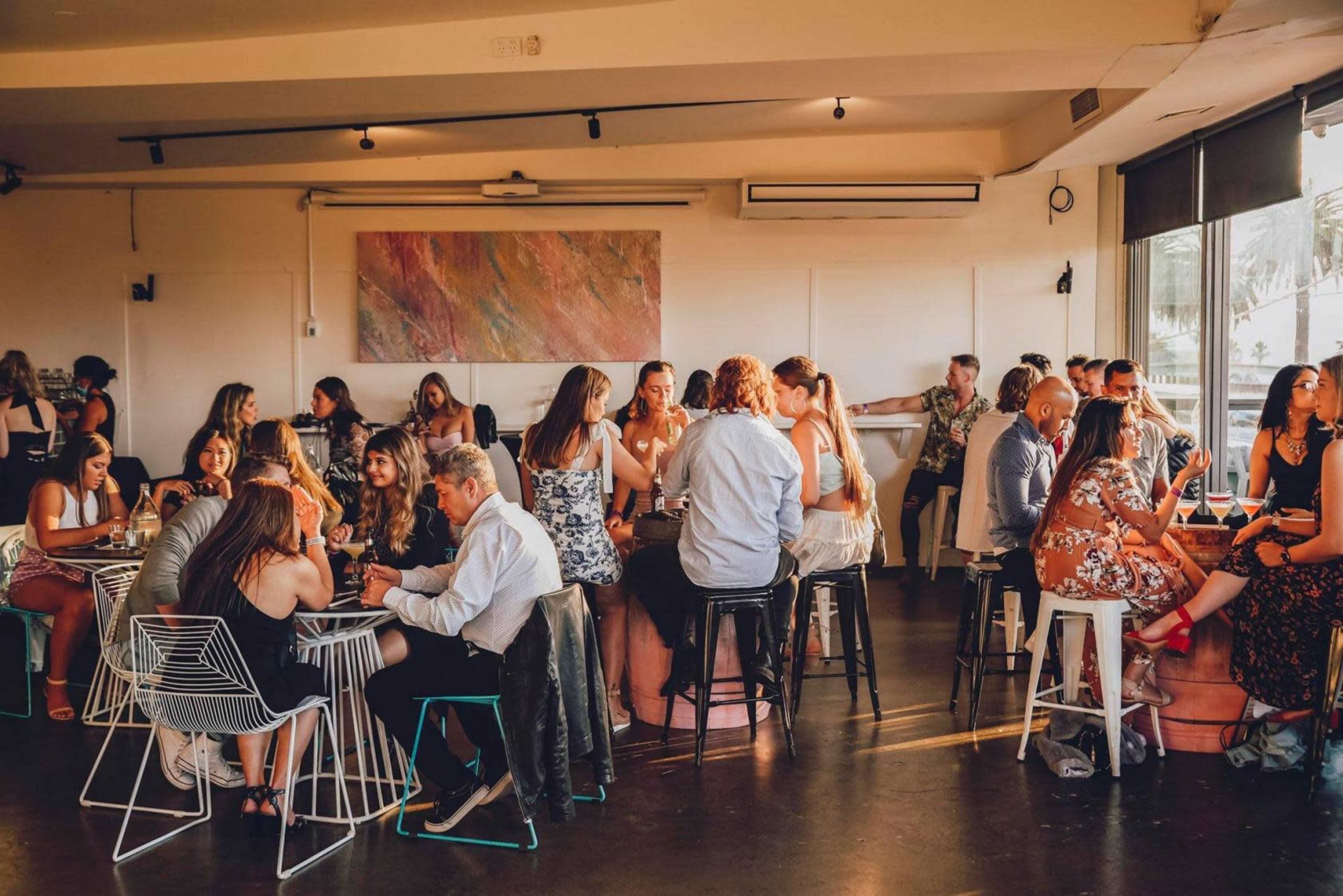 The image shows a lively restaurant scene with groups of people dining and socializing at various tables.
