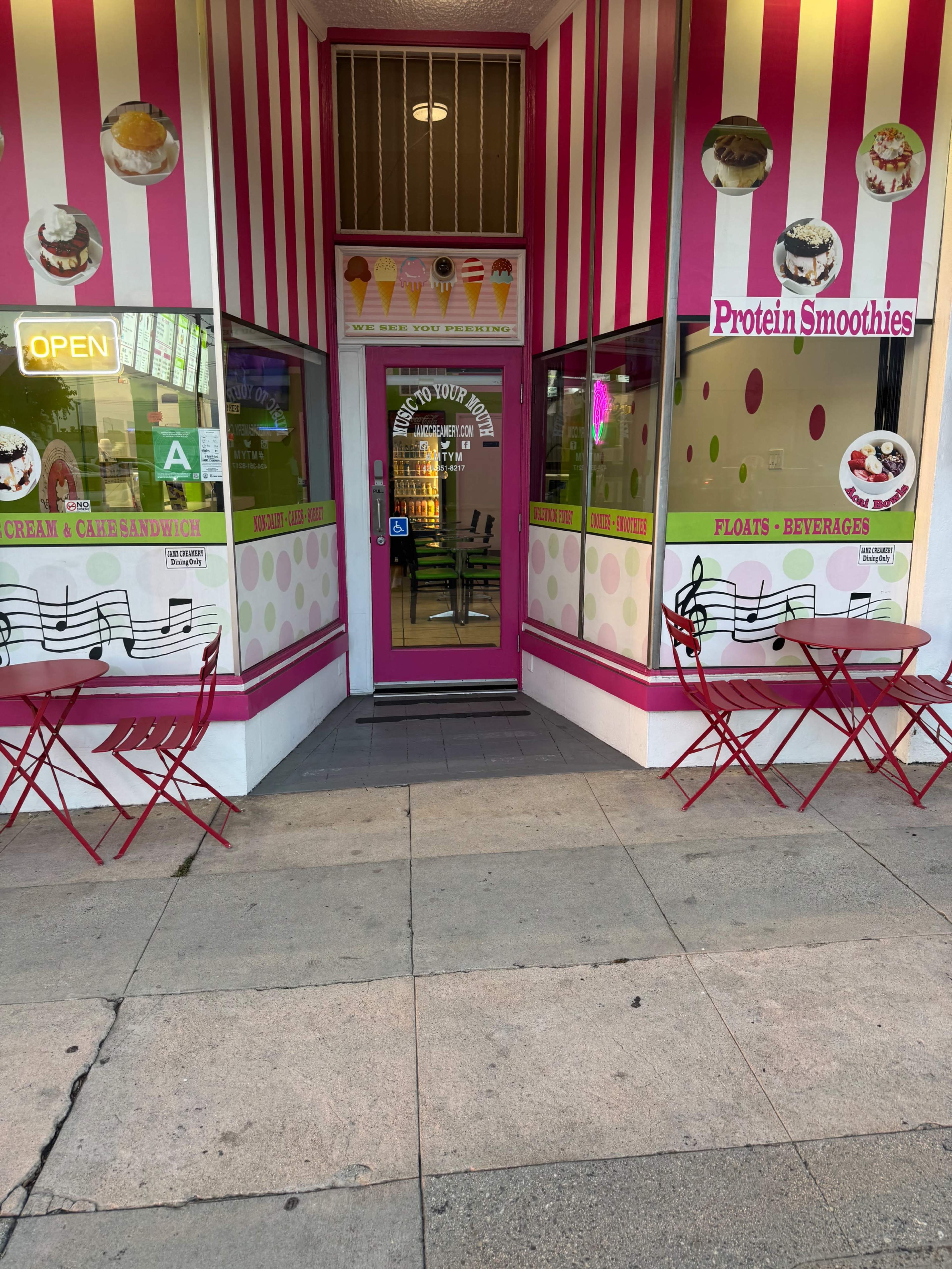 The image shows a storefront with pink and white striped walls featuring signs for protein smoothies, ice cream, and various beverages, along with small outdoor tables and chairs.