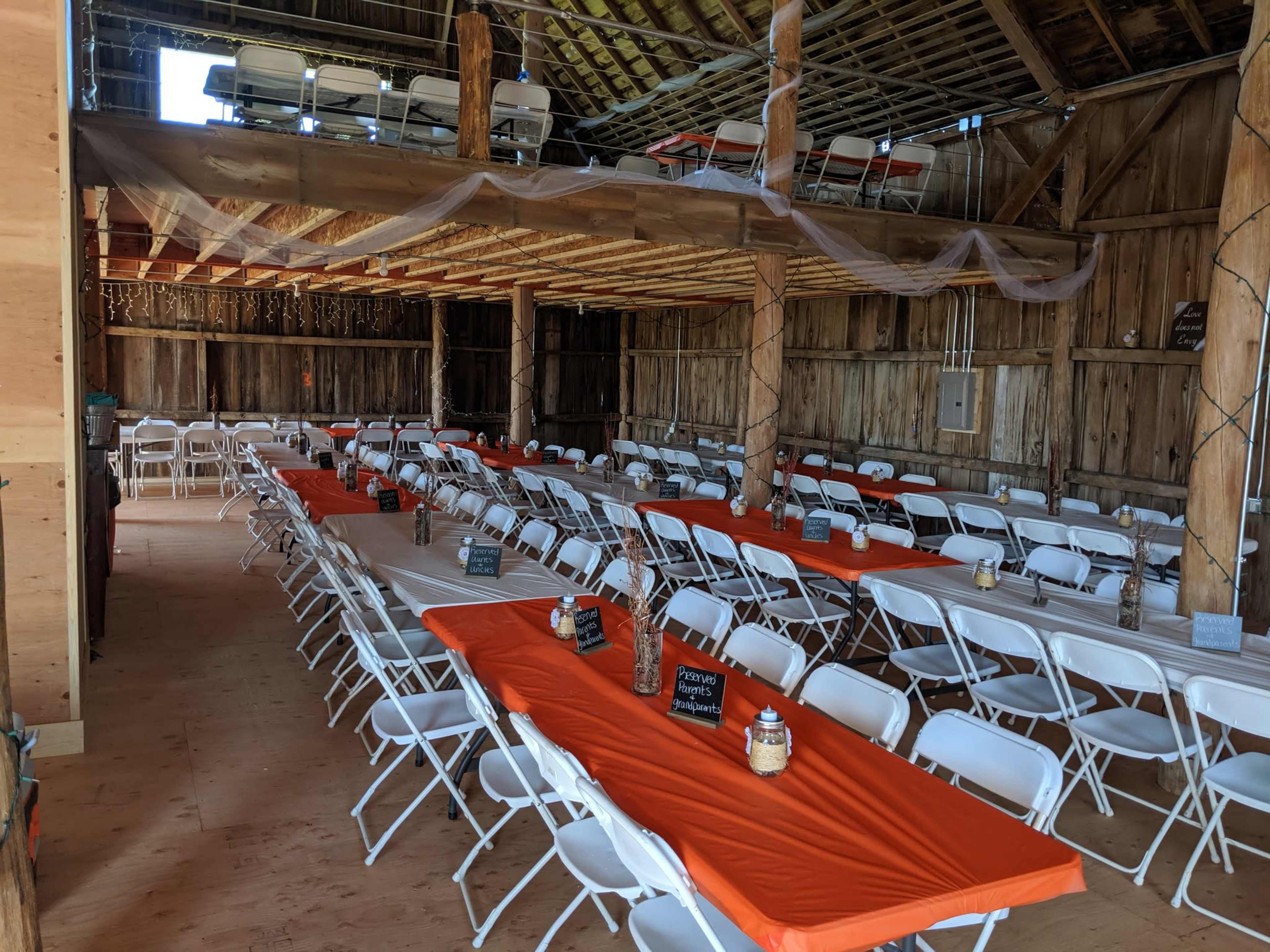 The image shows a large barn interior with rows of white folding tables covered in orange tablecloths and surrounded by white chairs, ready for an event.