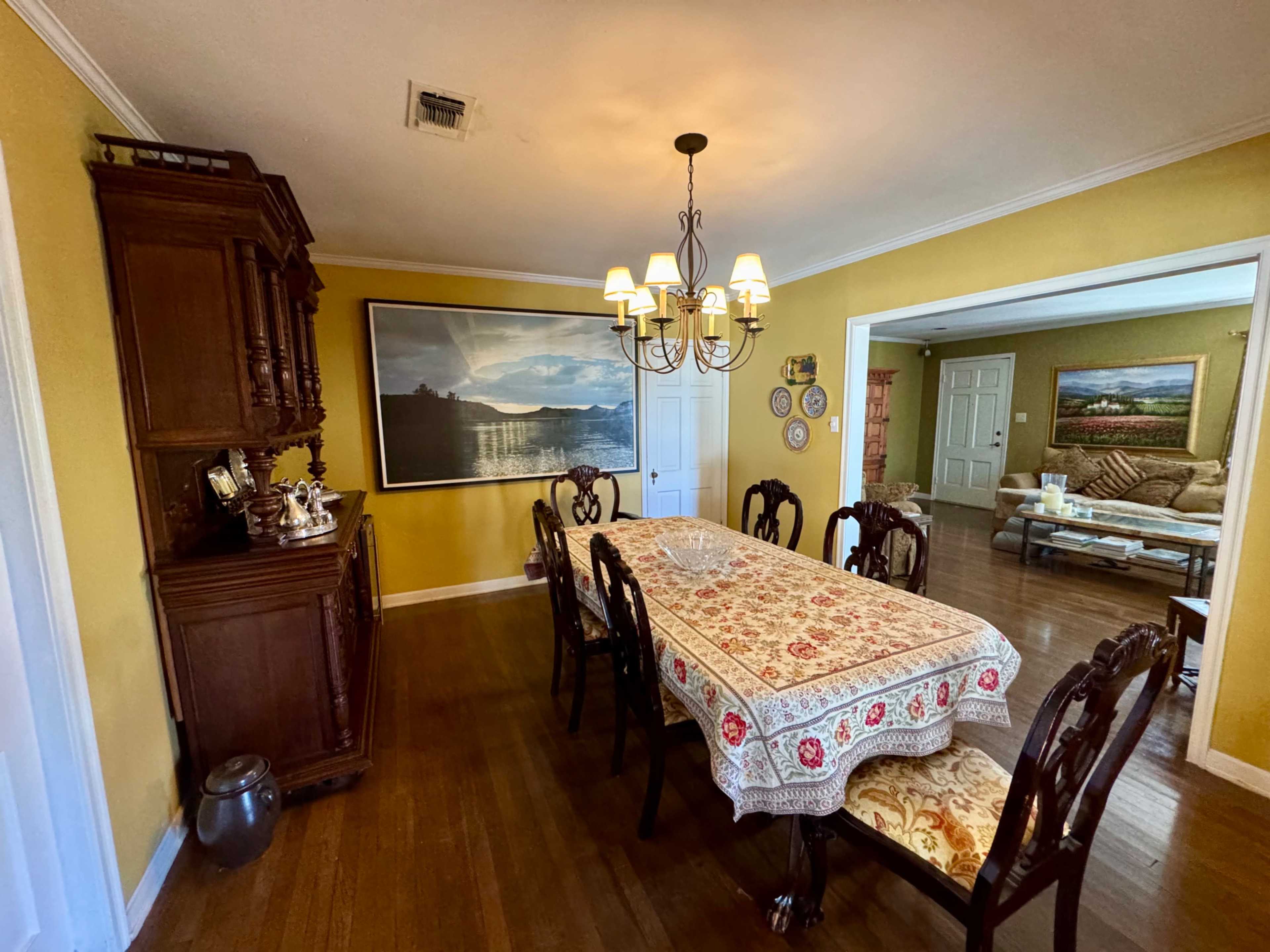 The image shows a spacious dining room with a long table covered by a decorative tablecloth, surrounded by wooden chairs, and a large painting on the wall.