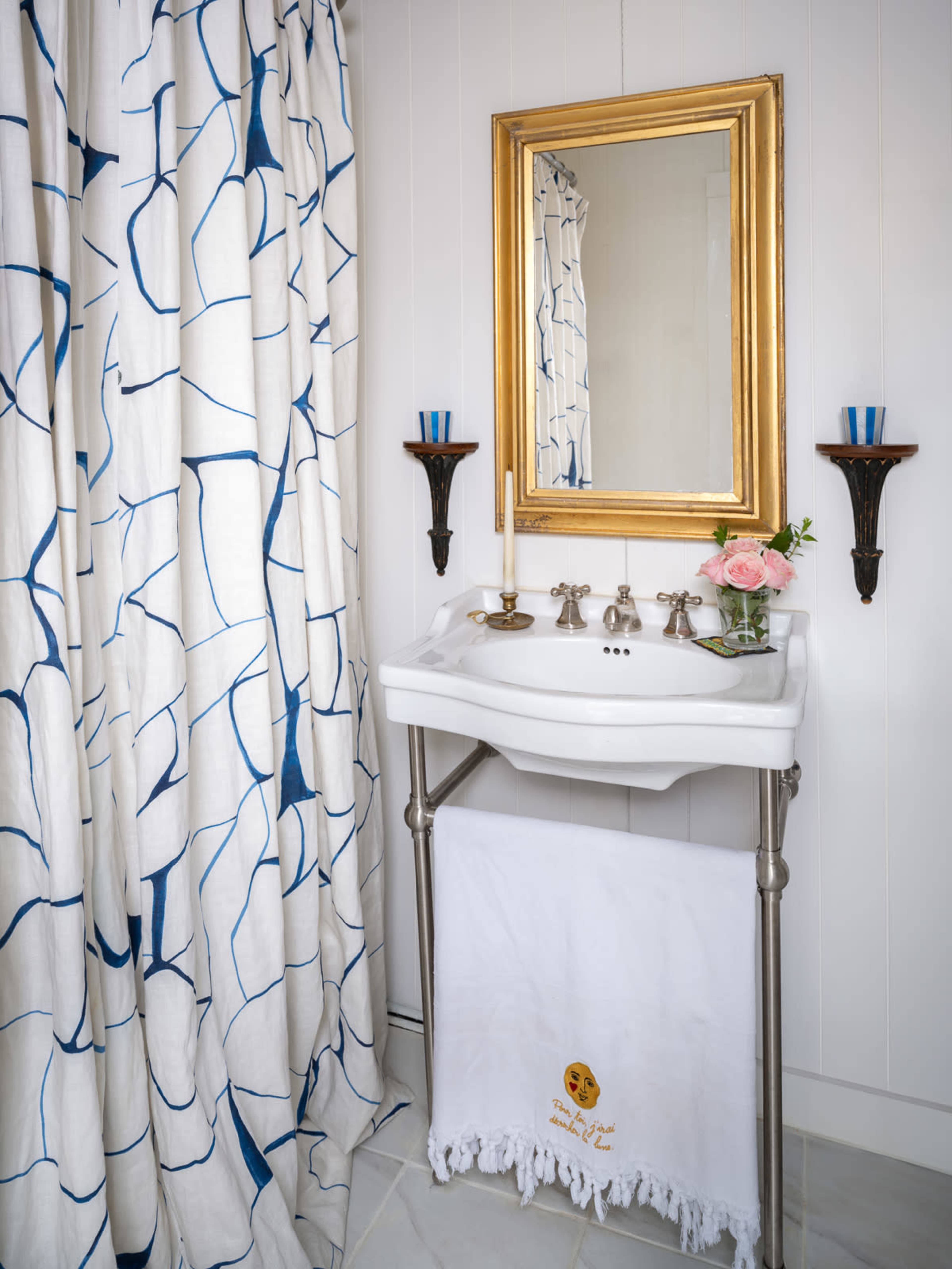A white basin with four faucet handles is situated beside a patterned shower curtain and a decorative mirror, accompanied by wall sconces holding candles.
