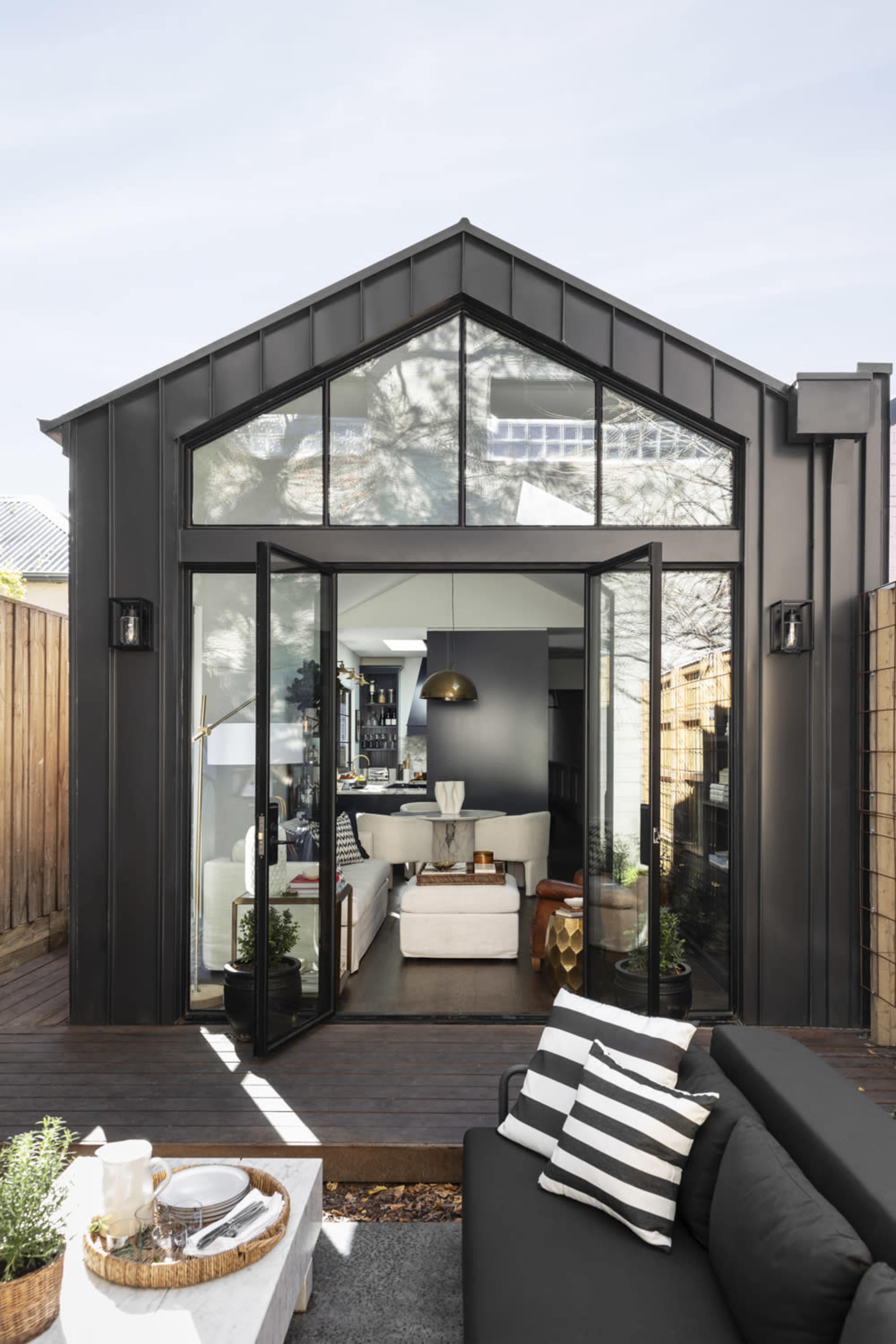 A modern black house with large glass doors opening to a living space, surrounded by a wooden deck and outdoor seating.