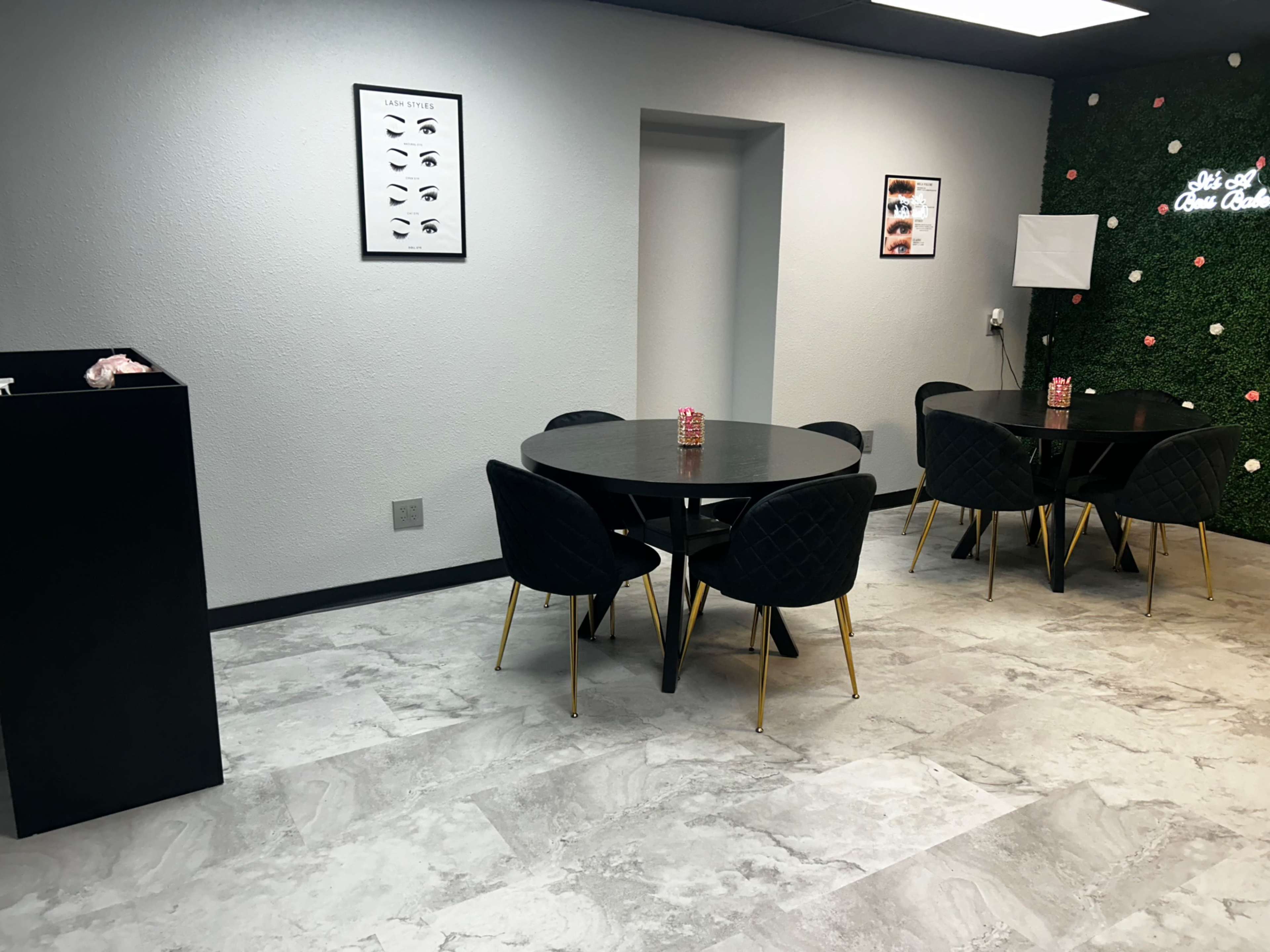 The image shows a modern, minimalist waiting area with two circular tables surrounded by black chairs, set against a light gray wall and a greenery accent wall.