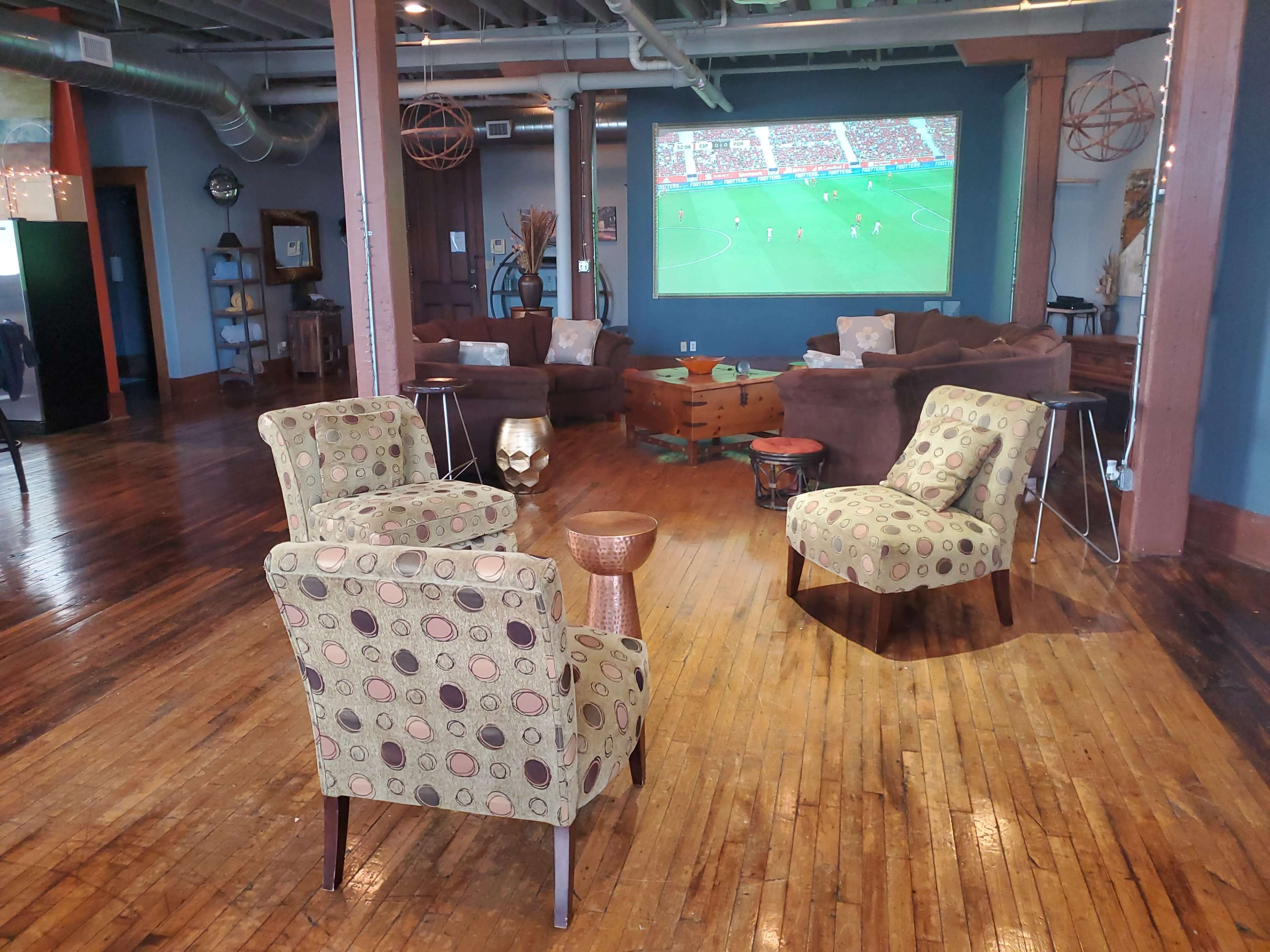 A spacious room with wooden floors, featuring three patterned chairs and a small table, while a large screen displays a soccer match in the background.