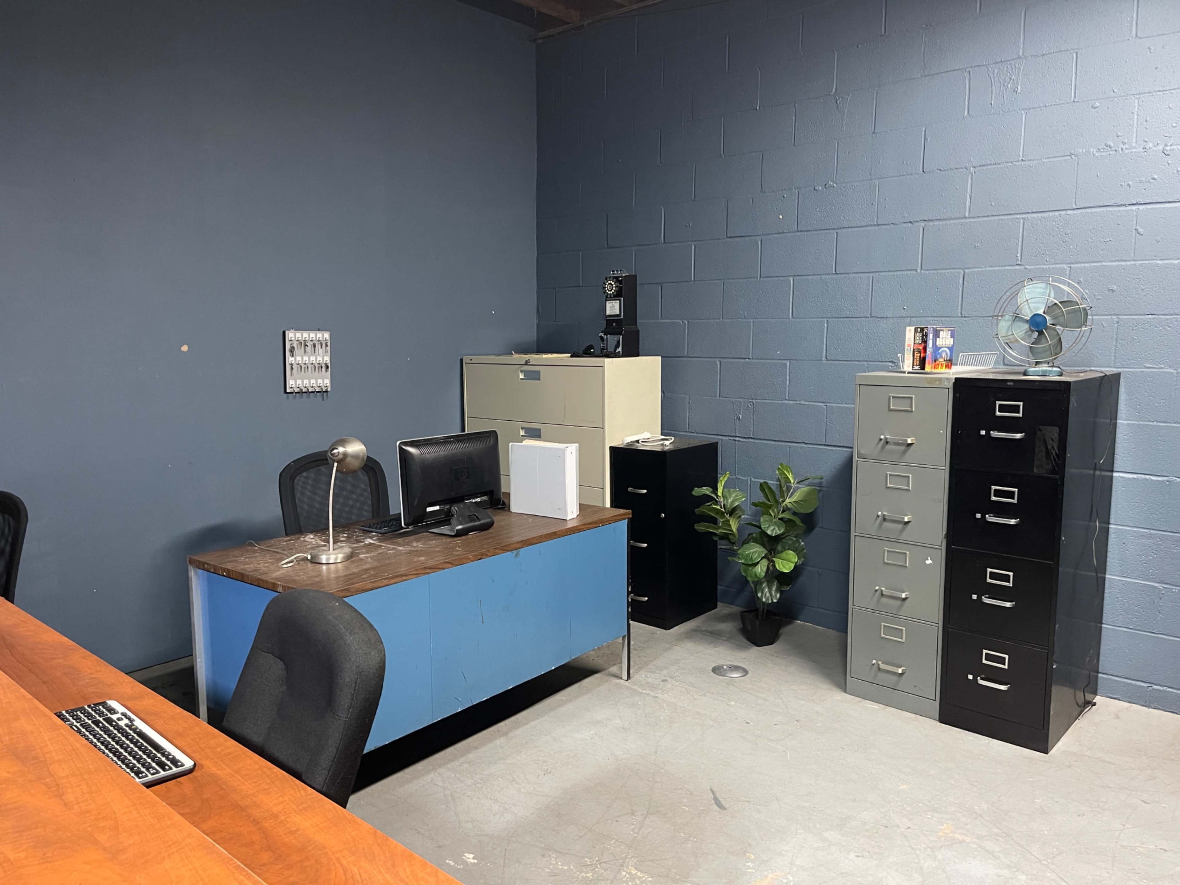 The image shows a simple office space with a blue desk holding a computer, black and gray filing cabinets, and a small plant in the corner.