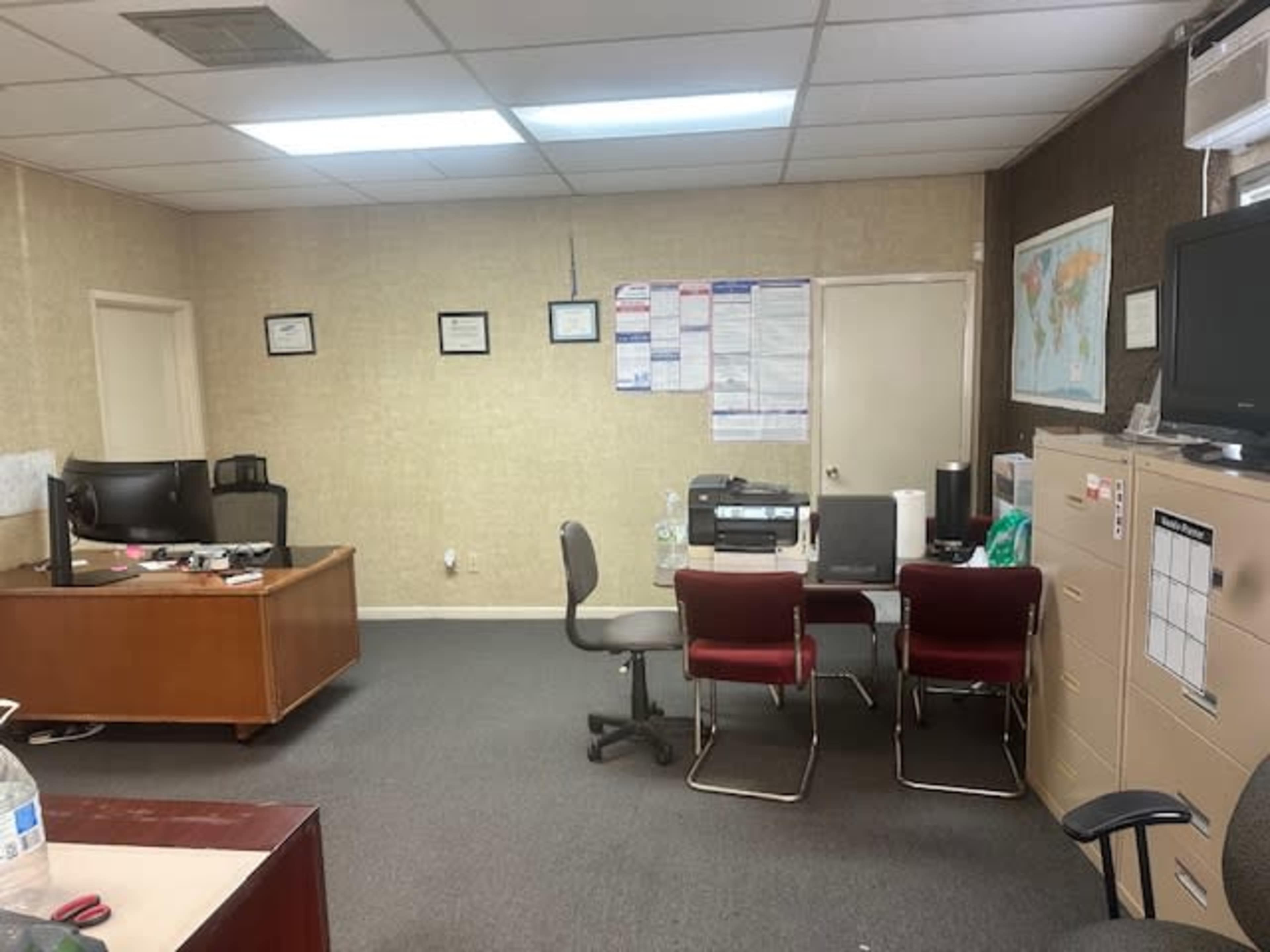 The image shows an office space with a desk, ergonomic chairs, a printer, and a wall displaying certificates and a map.