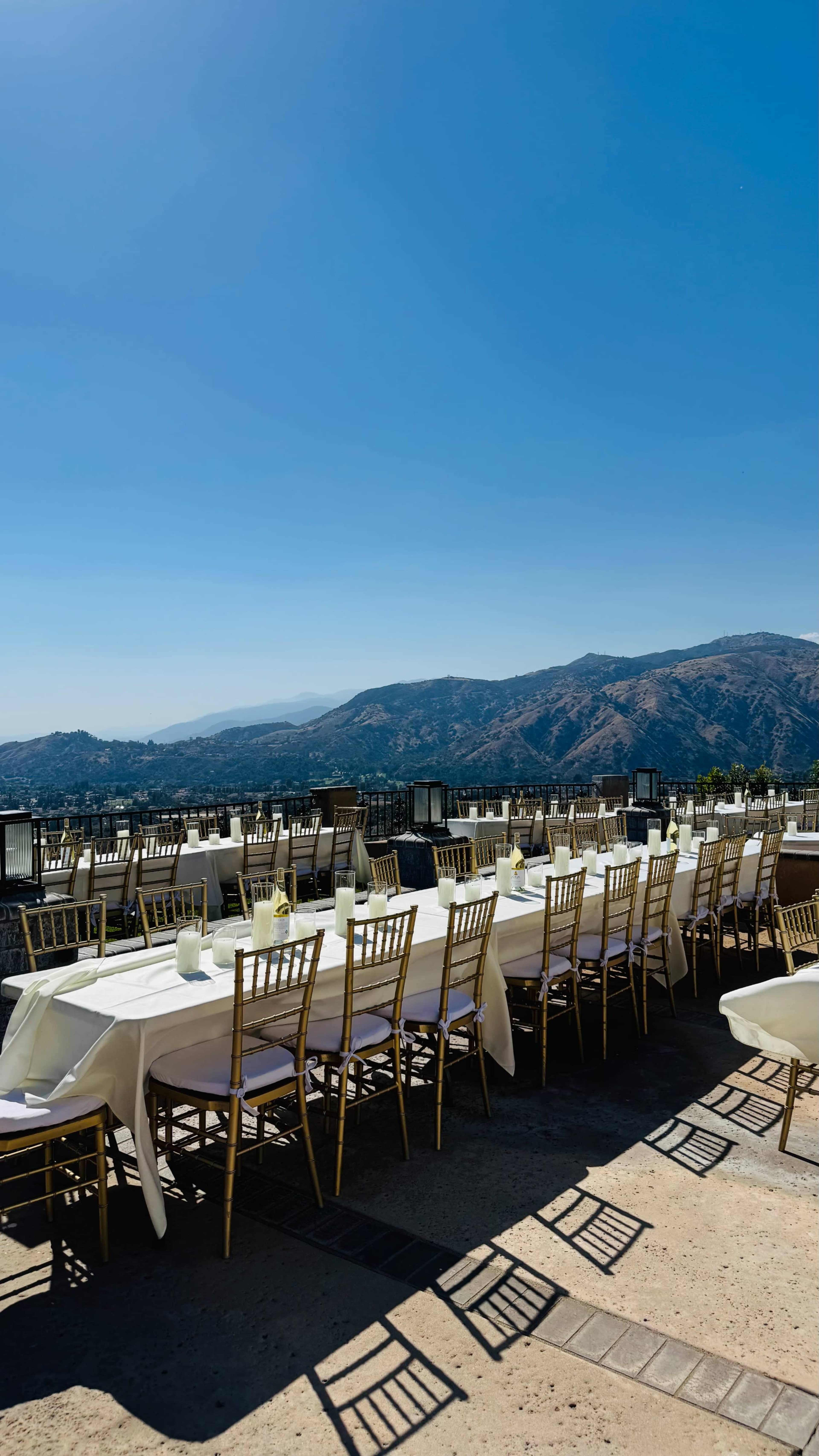 A long table set for dining with golden chairs overlooks a mountainous landscape under a clear blue sky.