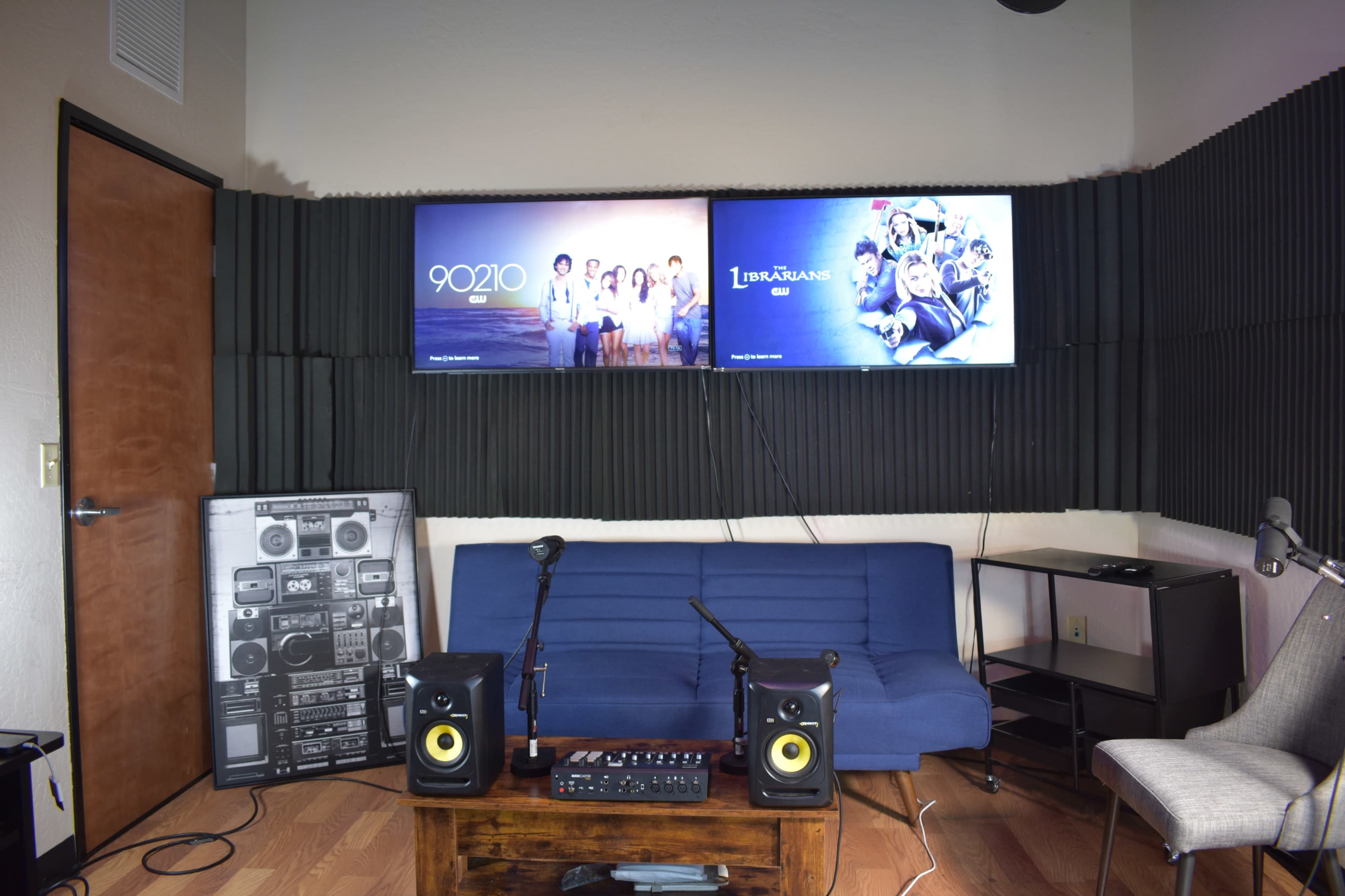A cozy room with two mounted television screens displaying shows, a blue couch, speakers on a wooden table, and soundproofing panels on the walls.