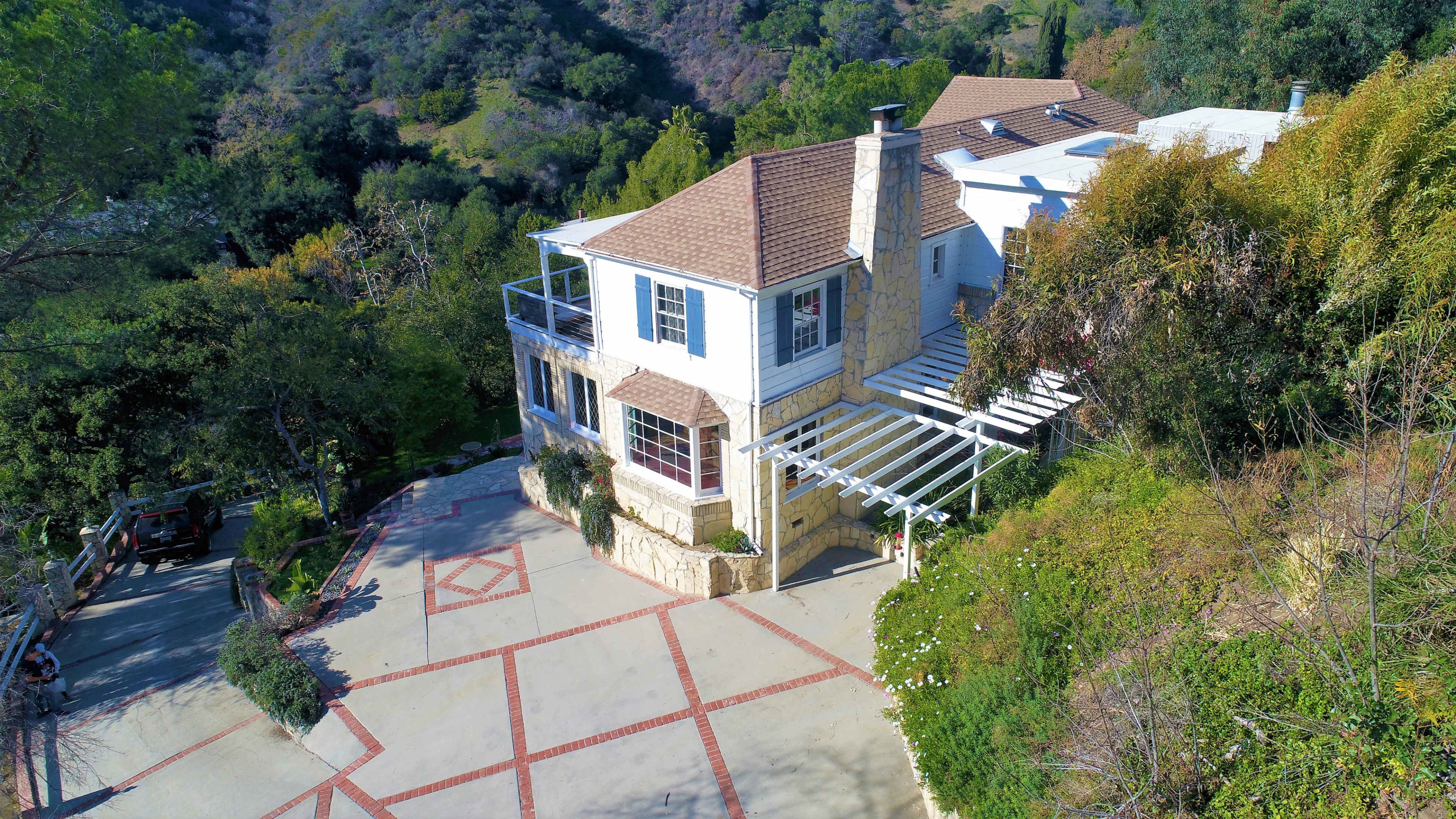 A two-story house with a stone exterior and a sloped roof is situated on a hillside, surrounded by greenery and a paved driveway.