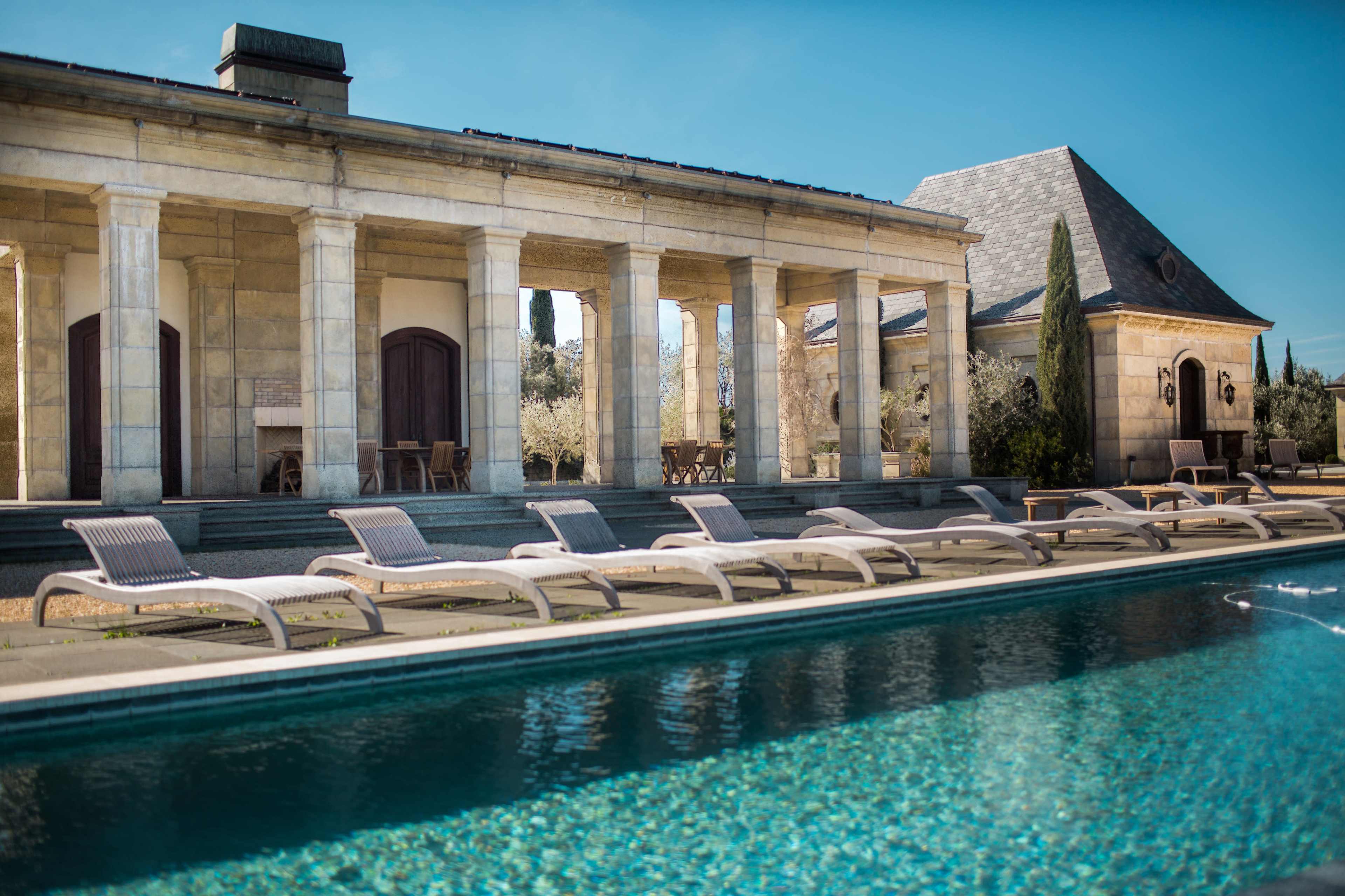 A serene outdoor swimming pool lined with lounge chairs in front of a stone building with columns and a roofed area.