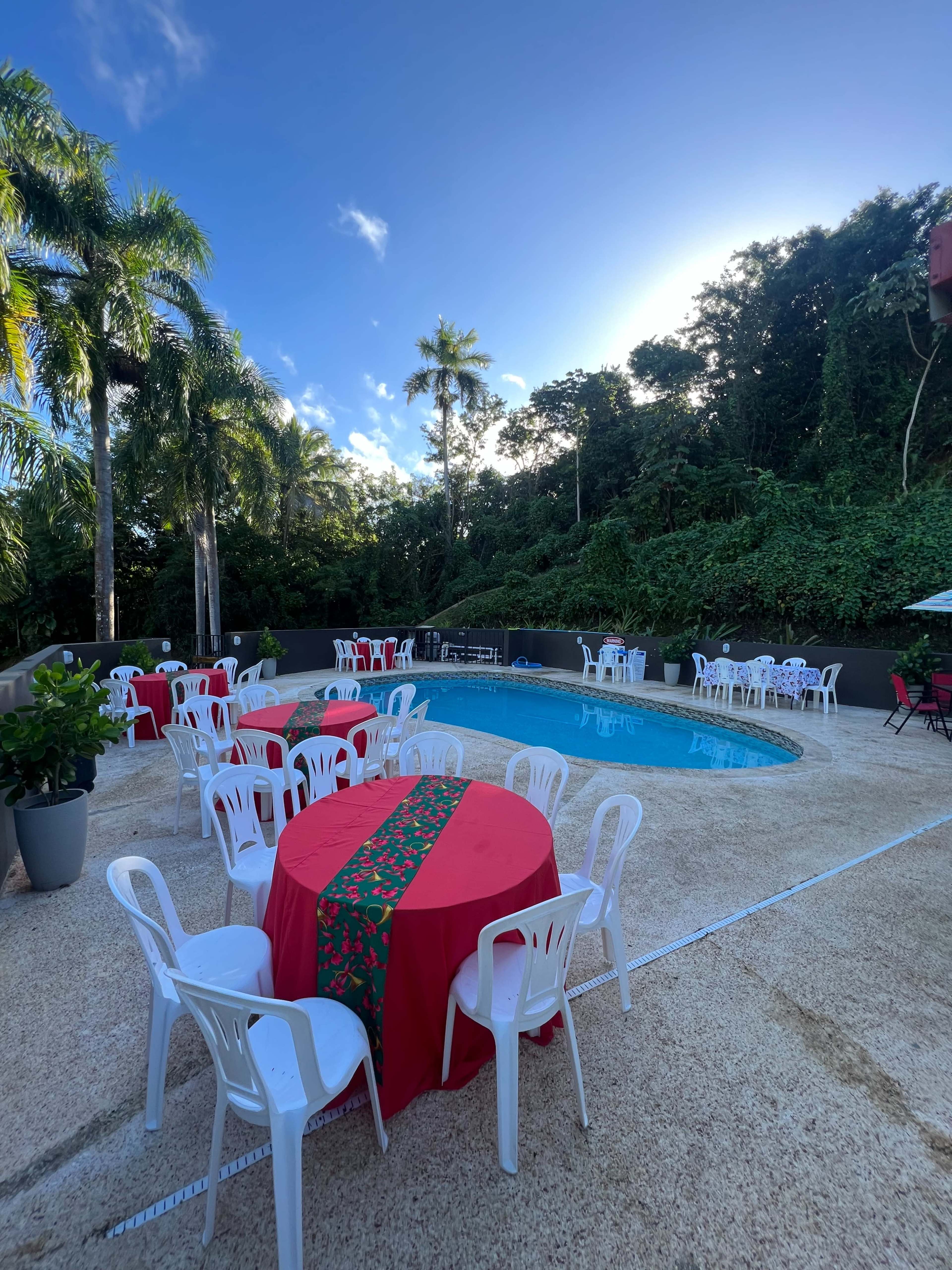 The image shows a pool area with several red tablecloth-covered tables and white chairs set up near a swimming pool, surrounded by lush greenery and palm trees.