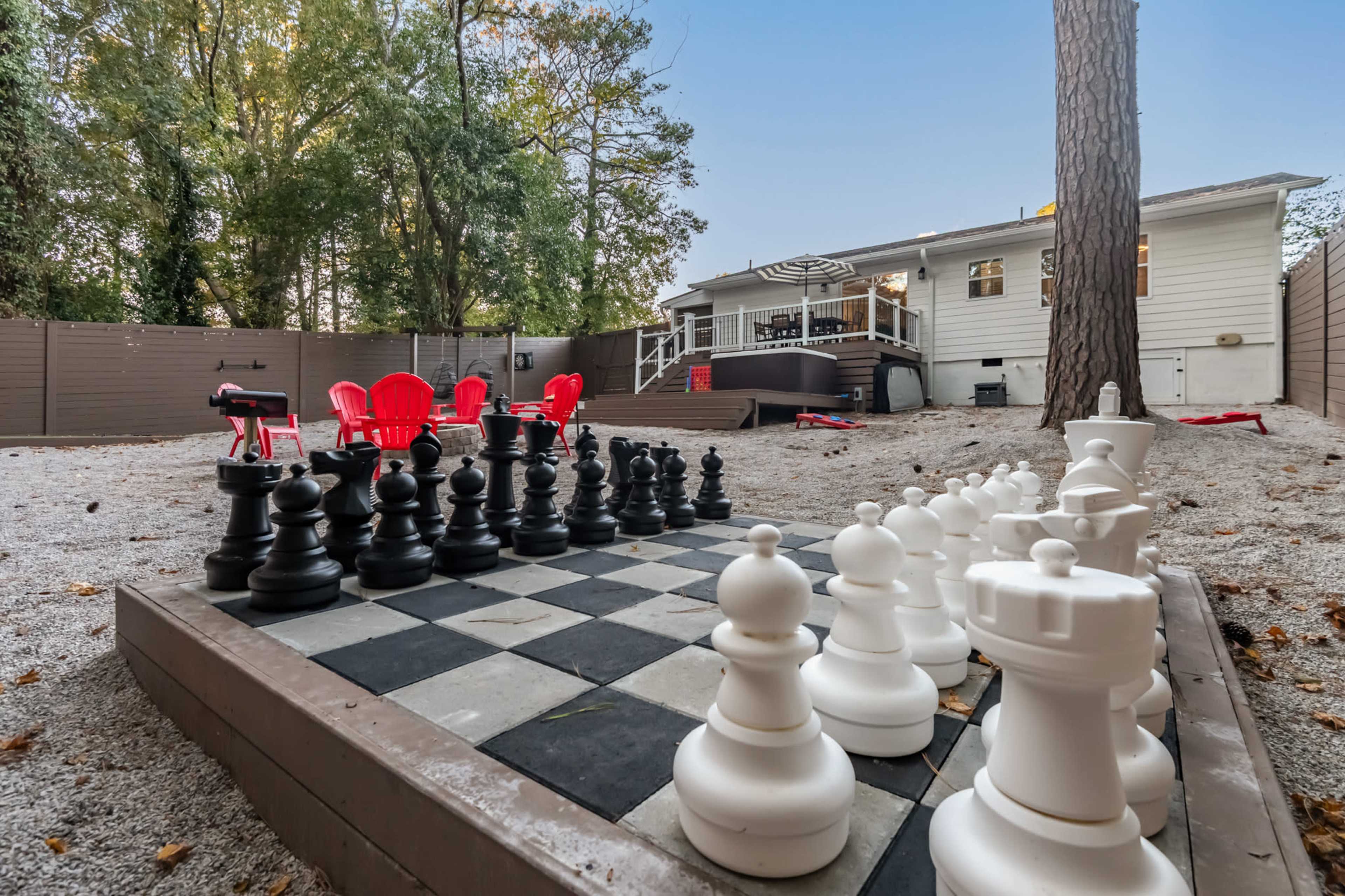 An oversized outdoor chessboard with black and white pieces is set in a gravel yard surrounded by trees, with a house and seating area in the background.