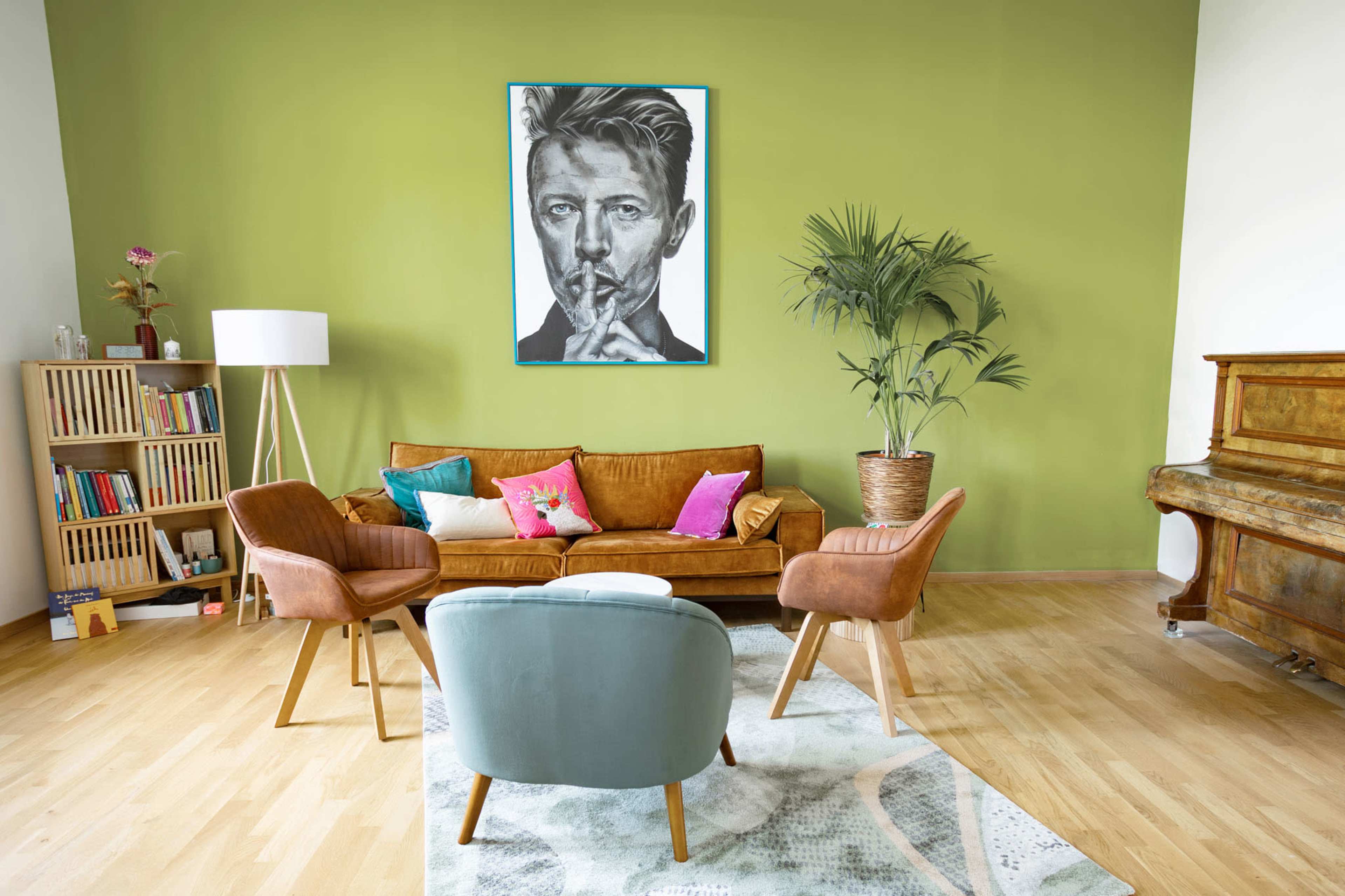 A modern living room features a green accent wall, a large artwork of a man, a brown velvet couch, armchairs, a patterned rug, and a wooden piano.