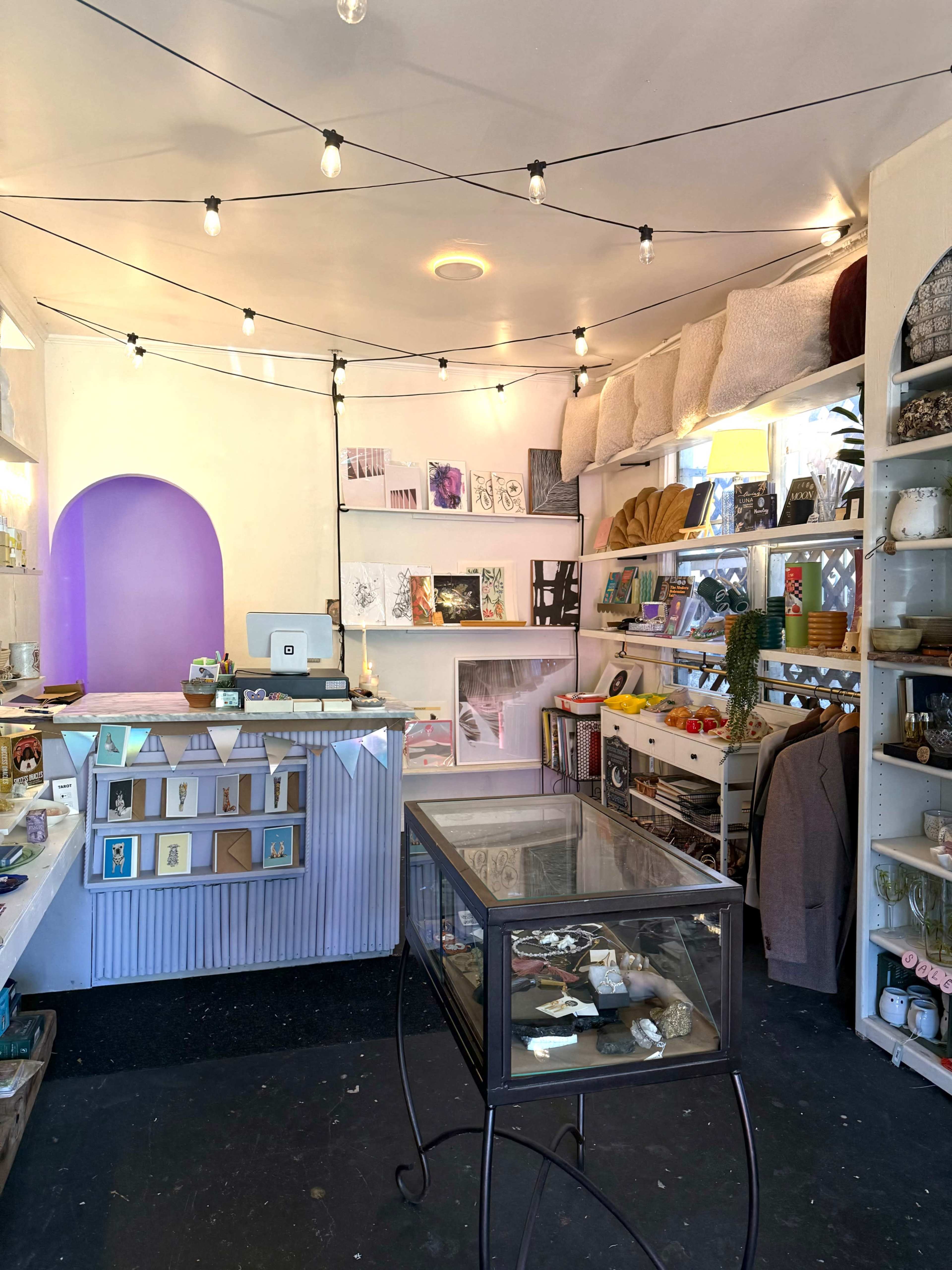 The image shows a small shop interior with a checkout counter, display shelves filled with various items, and decorative string lights overhead.