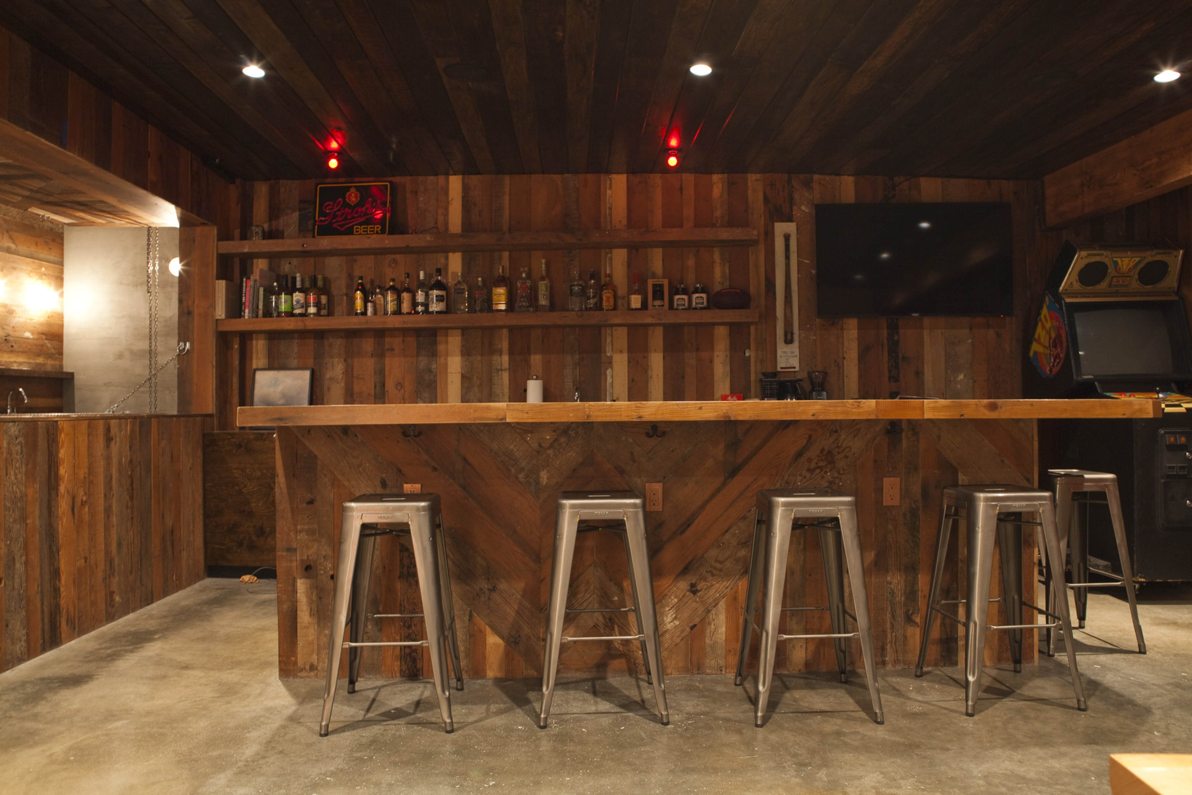 A rustic wooden bar with metal stools, a shelf of liquor bottles in the background, and a vintage arcade game in the corner.