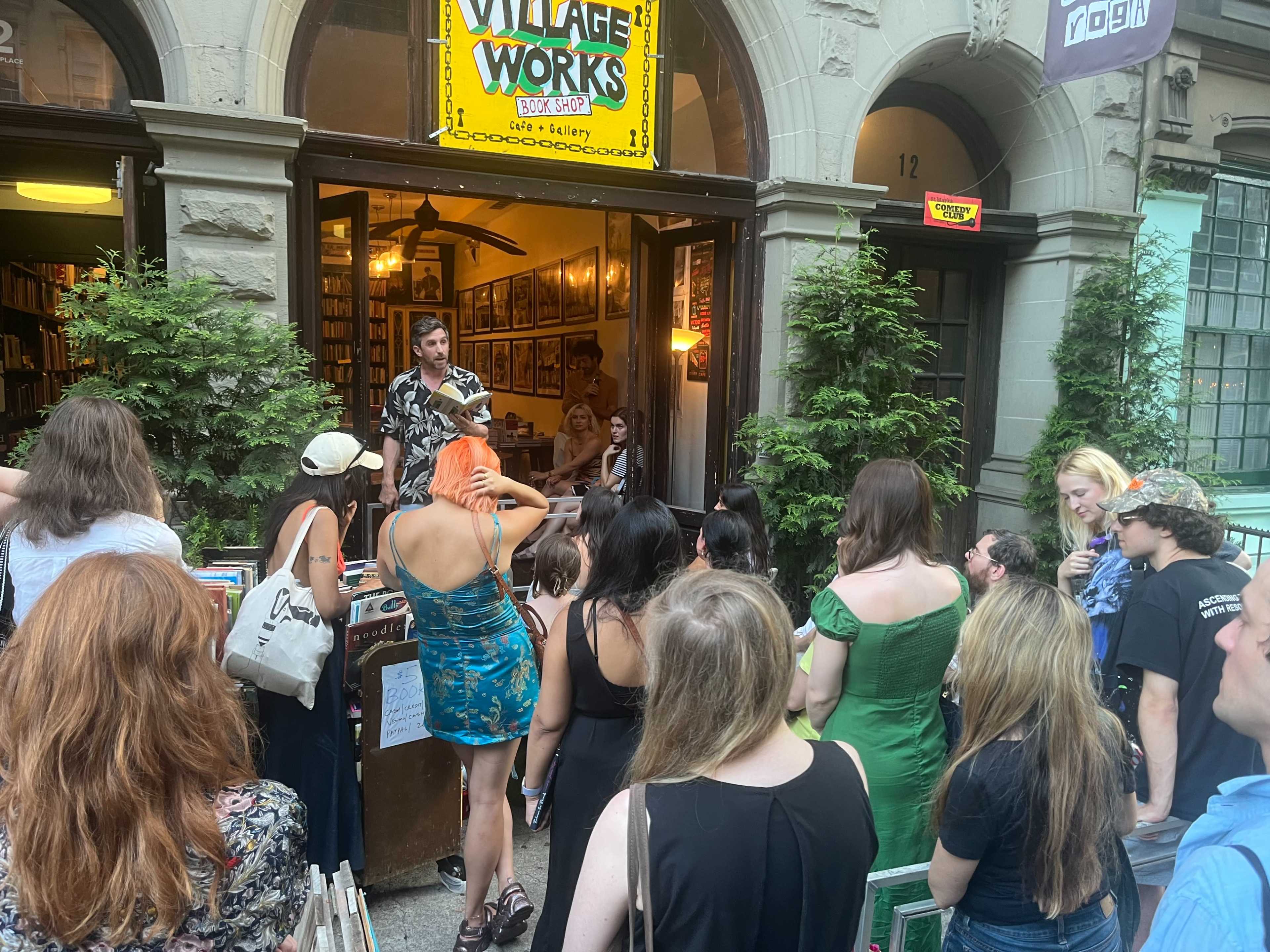 A crowd gathers outside a bookstore called Village Works, where a speaker addresses an audience while people browse books displayed at the entrance.