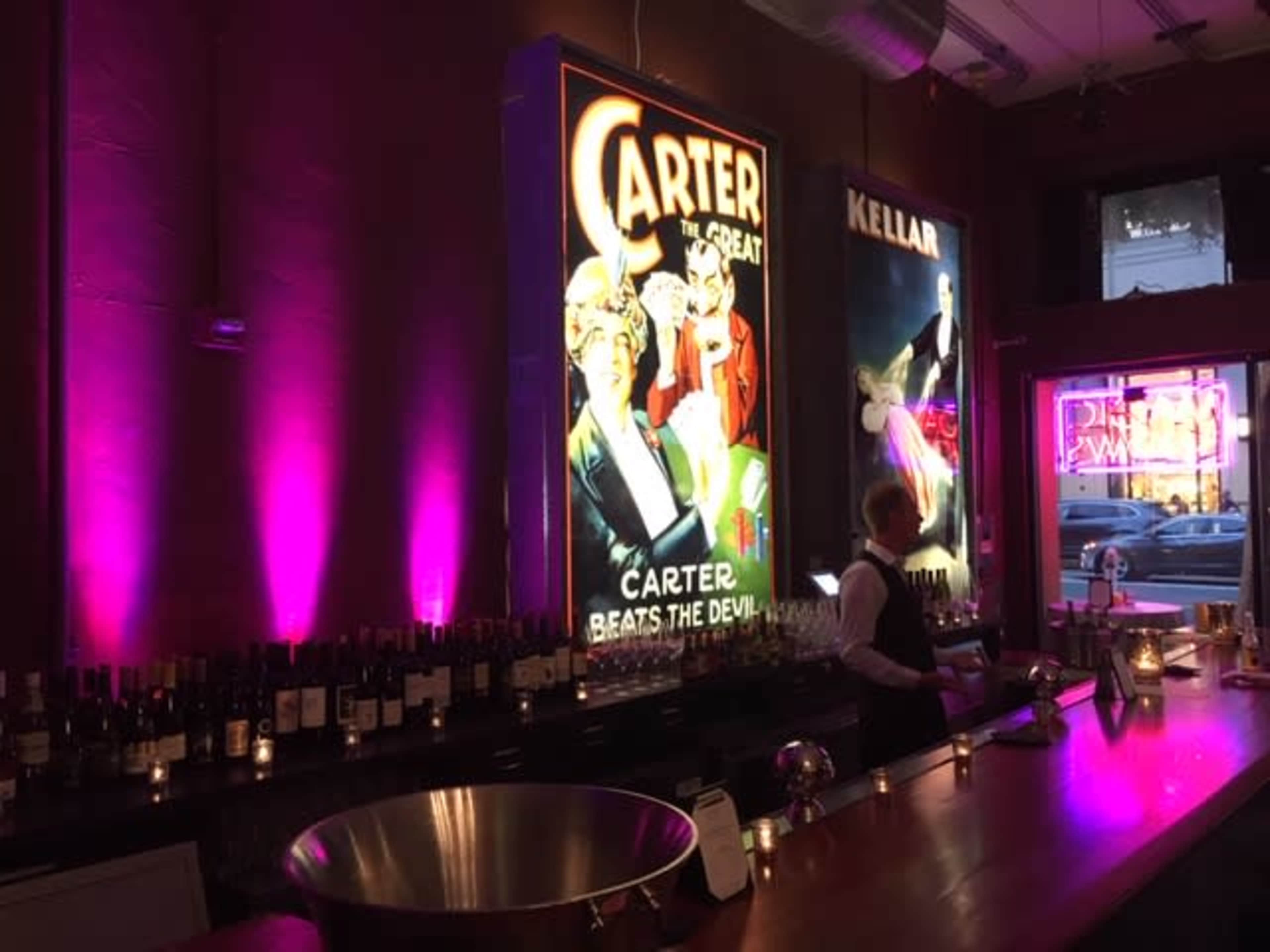 A bar area with illuminated vintage-style posters of magicians Carter and Kellar, against a backdrop of wine bottles and purple ambient lighting.